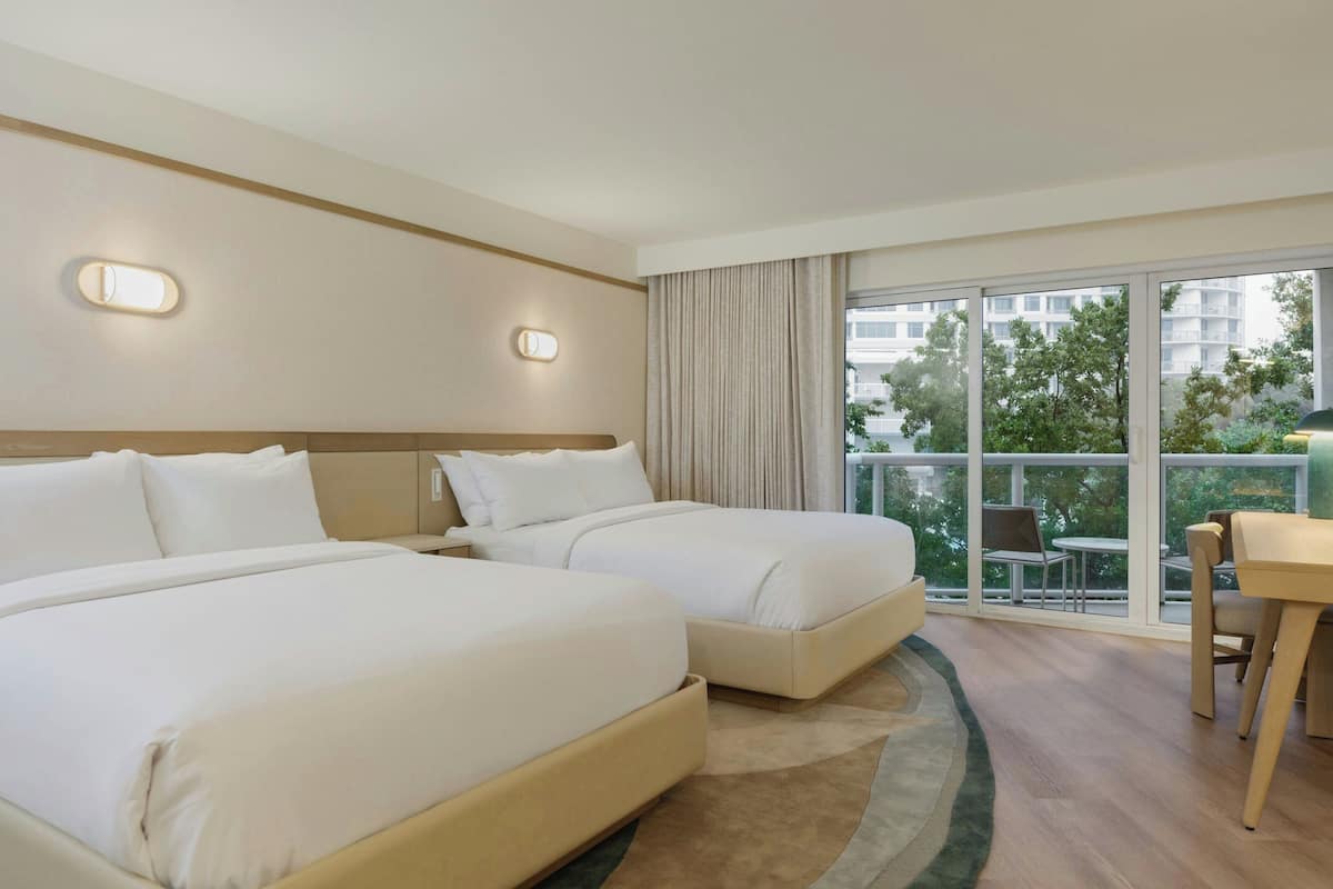 Une chambre d’hôtel avec deux lits, un bureau et un balcon offrant une vue sur les arbres et les bâtiments.