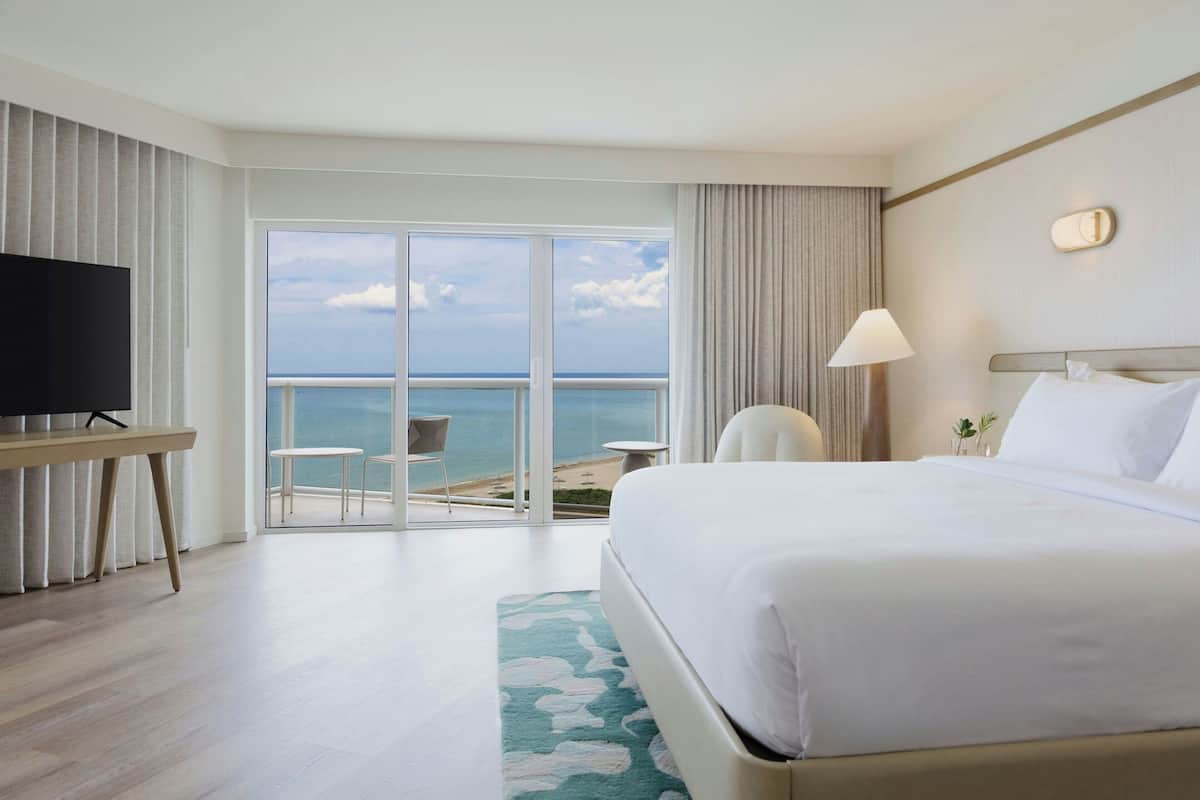 Une chambre d’hôtel avec un grand lit, une télévision, un bureau et une vue sur l’océan, accessible par une porte-fenêtre coulissante.