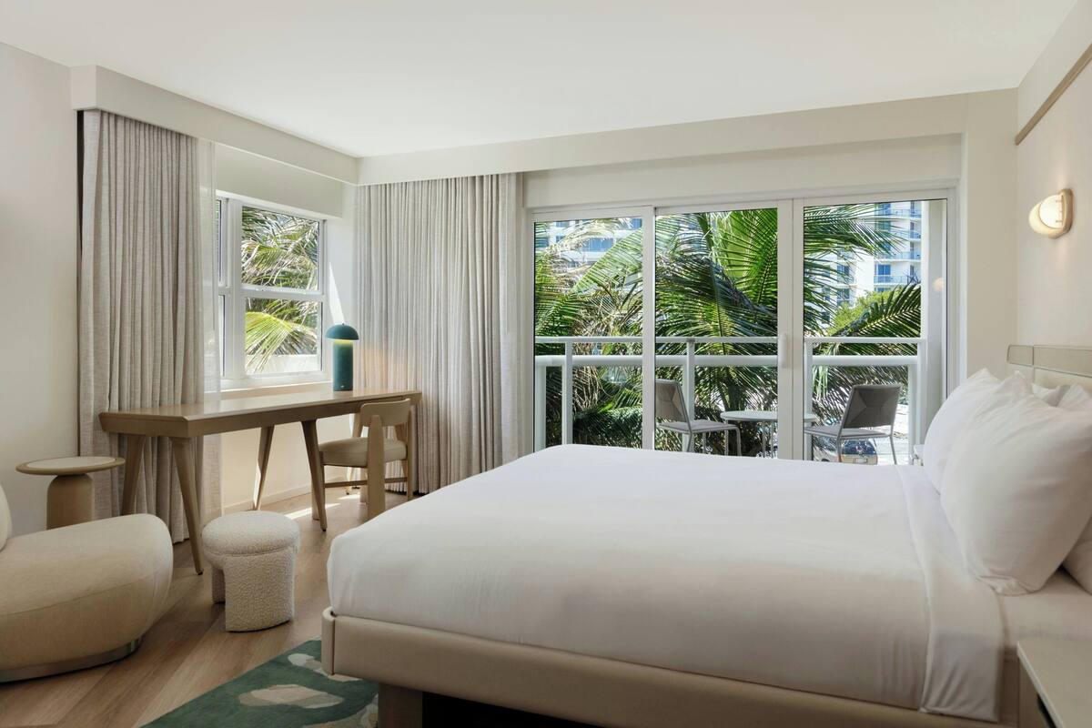 Une chambre d’hôtel avec un grand lit, un bureau, une chaise et une petite table. Il y a une fenêtre avec des rideaux, et de la fenêtre, on peut voir des palmiers et des immeubles.