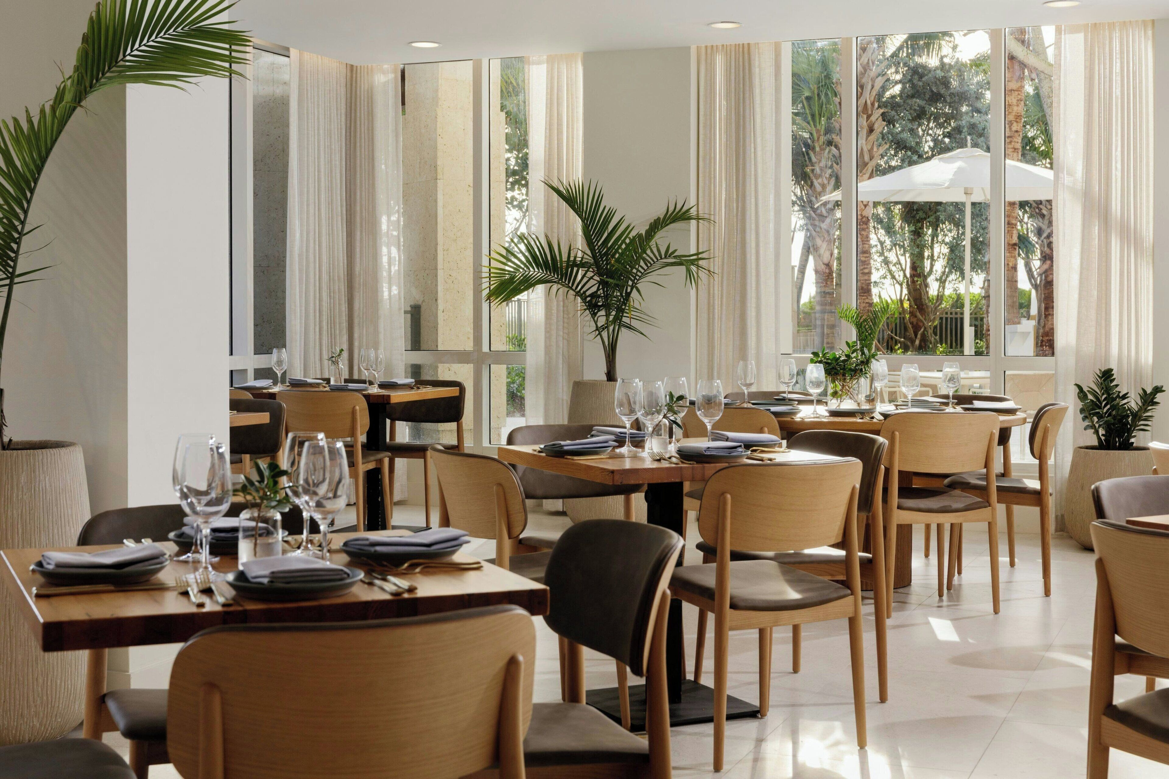 A restaurant with multiple tables and chairs, large windows with curtains, and indoor plants.