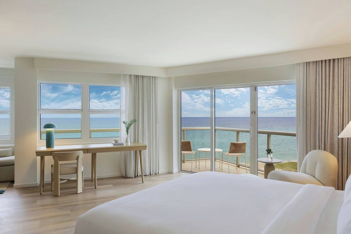 Une chambre d’hôtel dotée d’un grand lit, d’un bureau avec une chaise, d’une petite table avec une chaise et d’un balcon offrant une vue sur la mer.