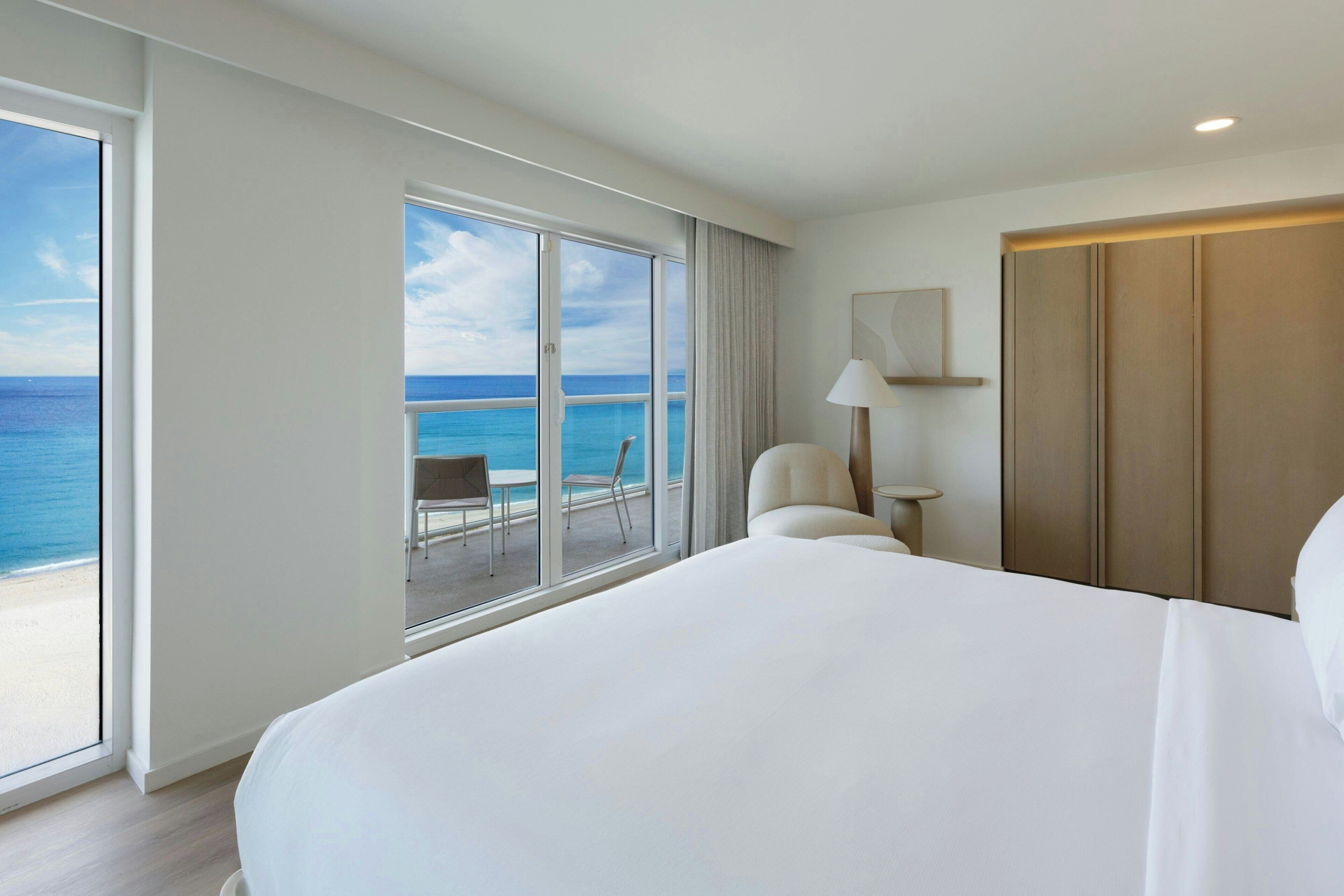 A hotel room with a large bed, a chair, a small table, and a view of the ocean through large windows.