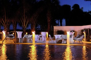 A poolside area with lounge chairs and tables, surrounded by palm trees.