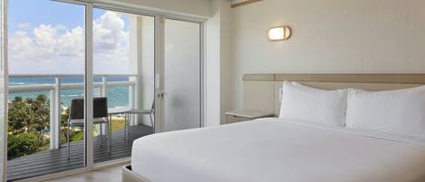 Habitación de hotel con una cama grande, vista al océano, un balcón con una silla y una mesita.