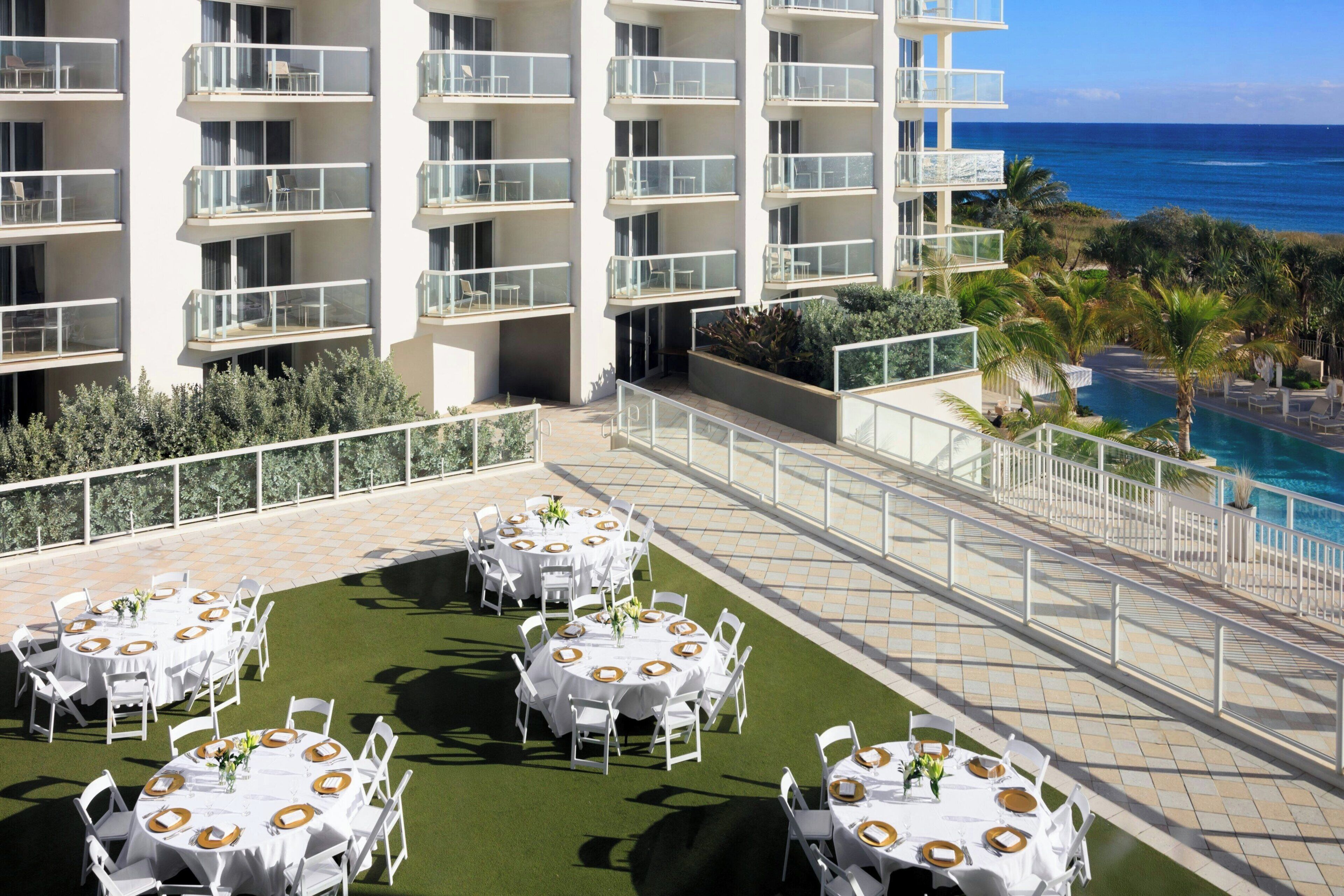 A rooftop dining area with round tables, white chairs, and a view of the ocean.