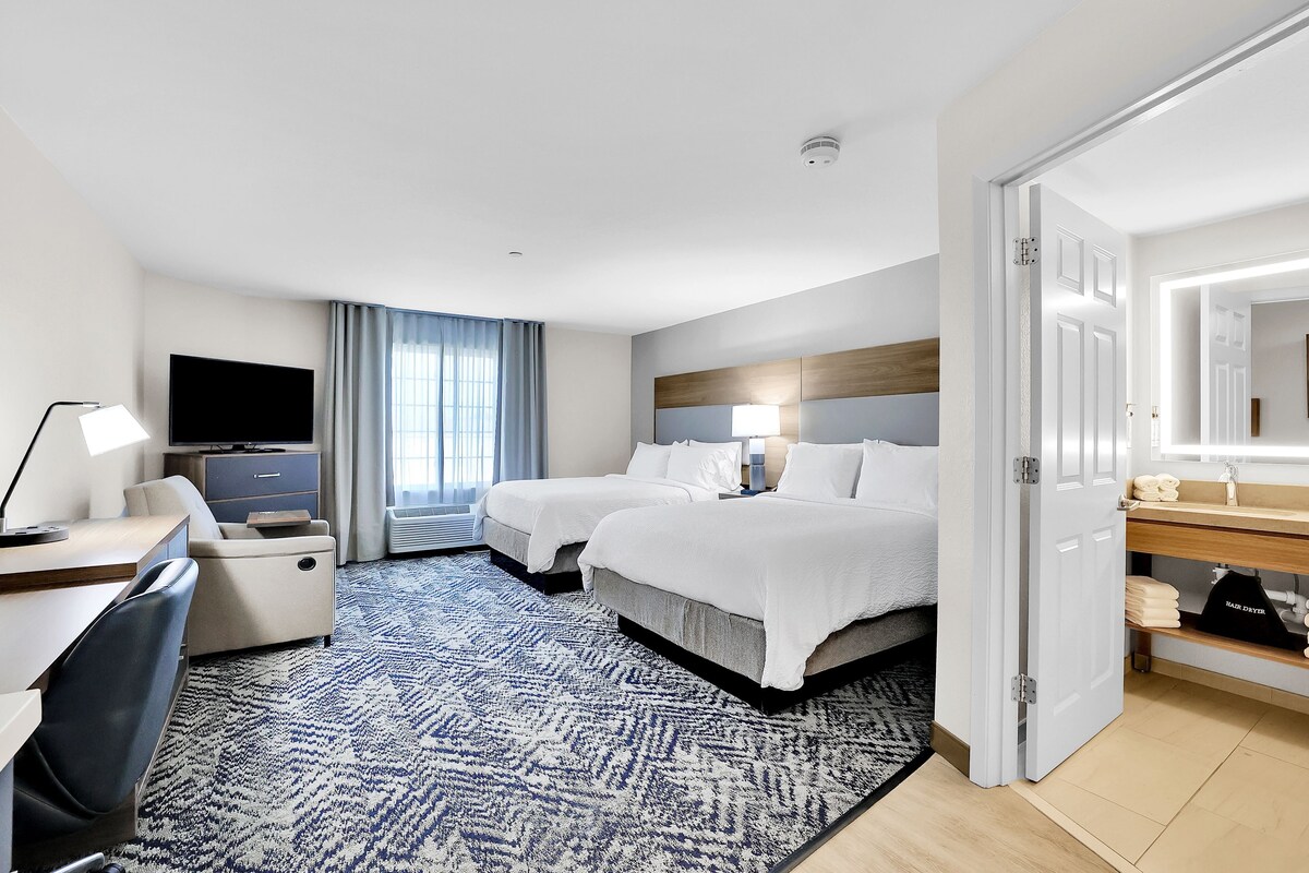 A hotel room with two beds, a desk, a TV, and a bathroom with a mirror and sink.