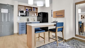 Una cocina moderna con encimera blanca, refrigerador negro y sillas azules. Hay un estante de madera sobre el lavabo y un espejo en la pared.