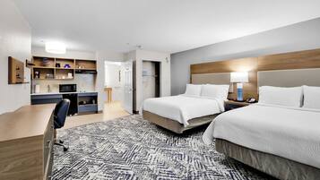 Una habitación de hotel con escritorio, dos camas, kitchenette y alfombra estampada.