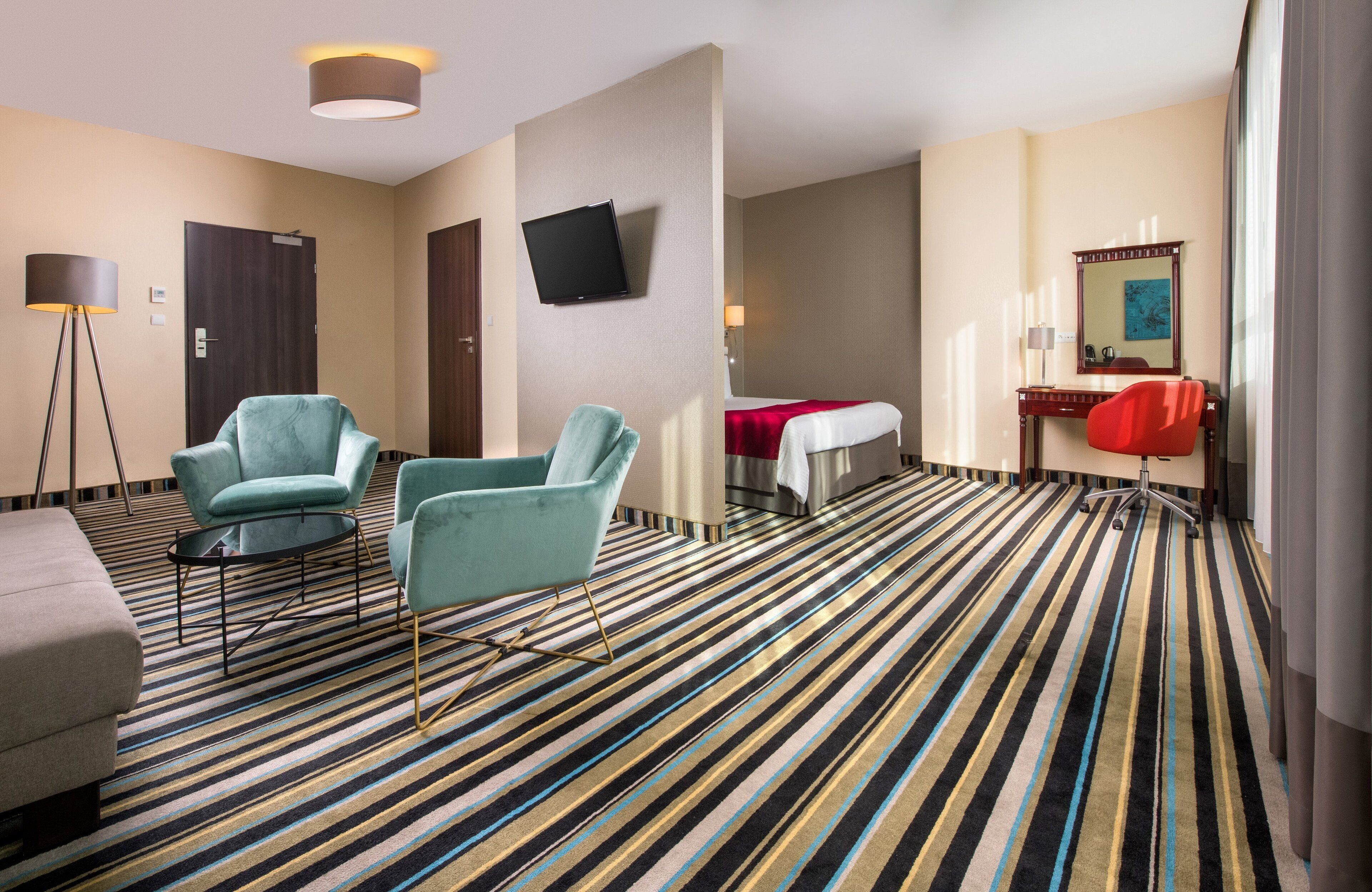 Ein Hotelzimmer mit Streifenmuster-Teppich, zwei Sesseln, einem Schreibtisch mit Stuhl, einem Bett, einem Fernseher, einem Spiegel und einer Stehlampe.
