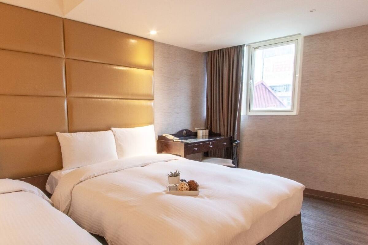 Ein Hotelzimmer mit einem großen Bett, einem Nachttisch, einer Lampe, einem Fenster mit Vorhängen und Blick auf ein Gebäude.