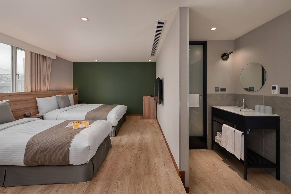 Ein Hotelzimmer mit zwei Betten, einem großen Fenster, einem Badezimmer mit Waschbecken und Spiegel sowie Blick auf die Stadt.