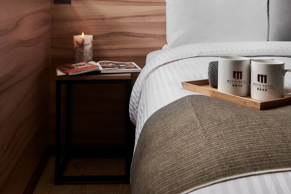 Ein ordentlich bezogenes Bett mit einem Holz-Kopfteil, ein Nachttisch mit einem Buch und einer Kerze sowie zwei Tassen auf einem Tablett.