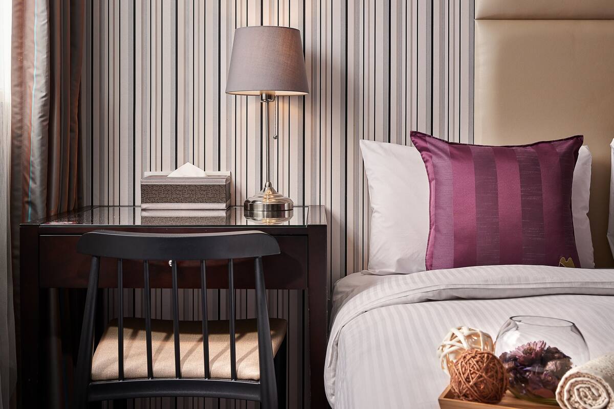 Ein Hotelzimmer mit gestreifter Tapete, einem Schreibtisch mit Lampe, einem Stuhl, einem Bett mit Kopfkissen und einem Tablett mit Dekoration.