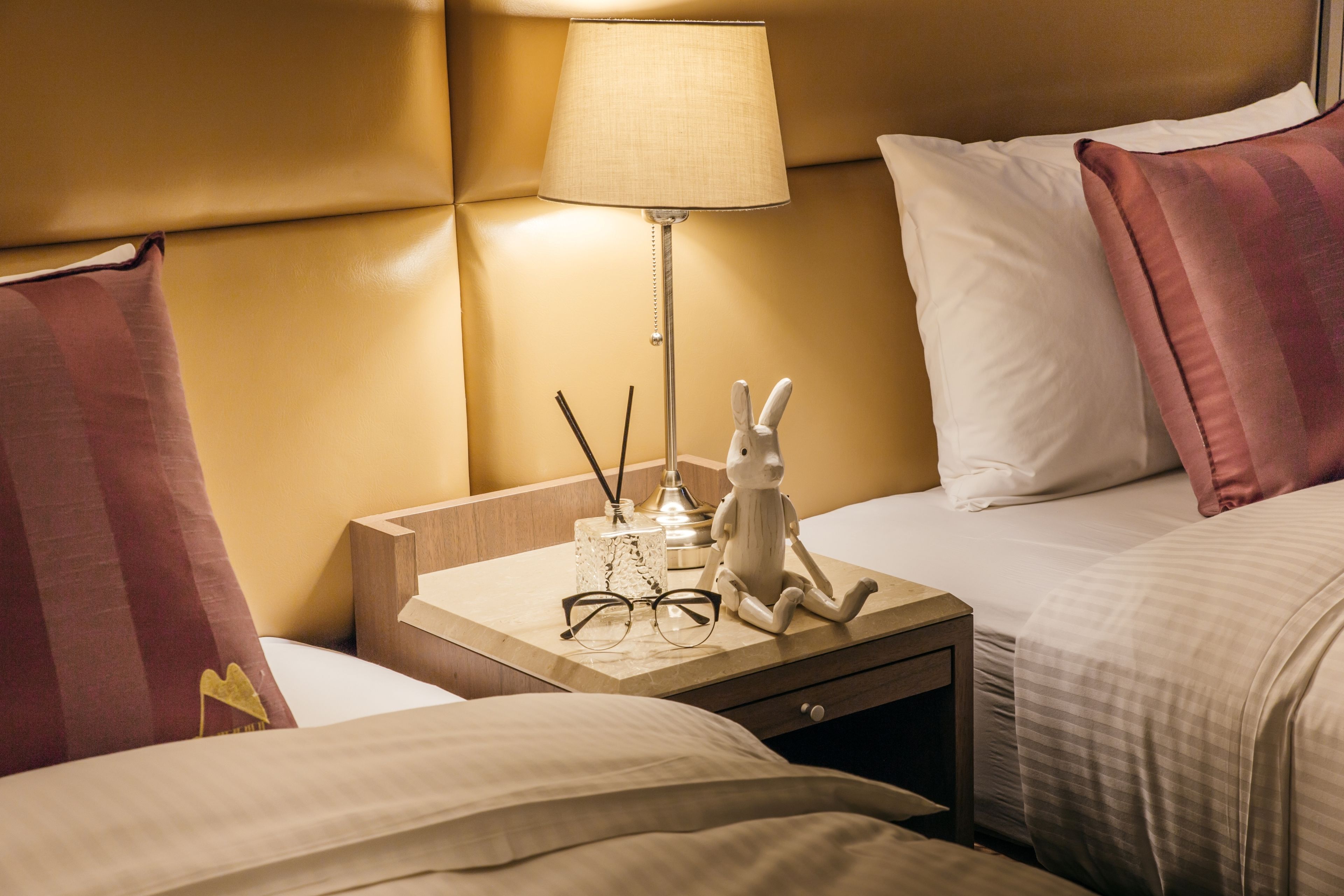 A hotel room with a bed, a nightstand with a lamp, a decorative bunny, and a pair of glasses.