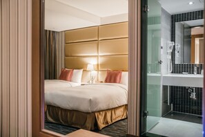 Hotelkamer met twee bedden, een groot gestoffeerd hoofdeinde, een nachtkastje met lamp en een douchecabine met glazen wanden.