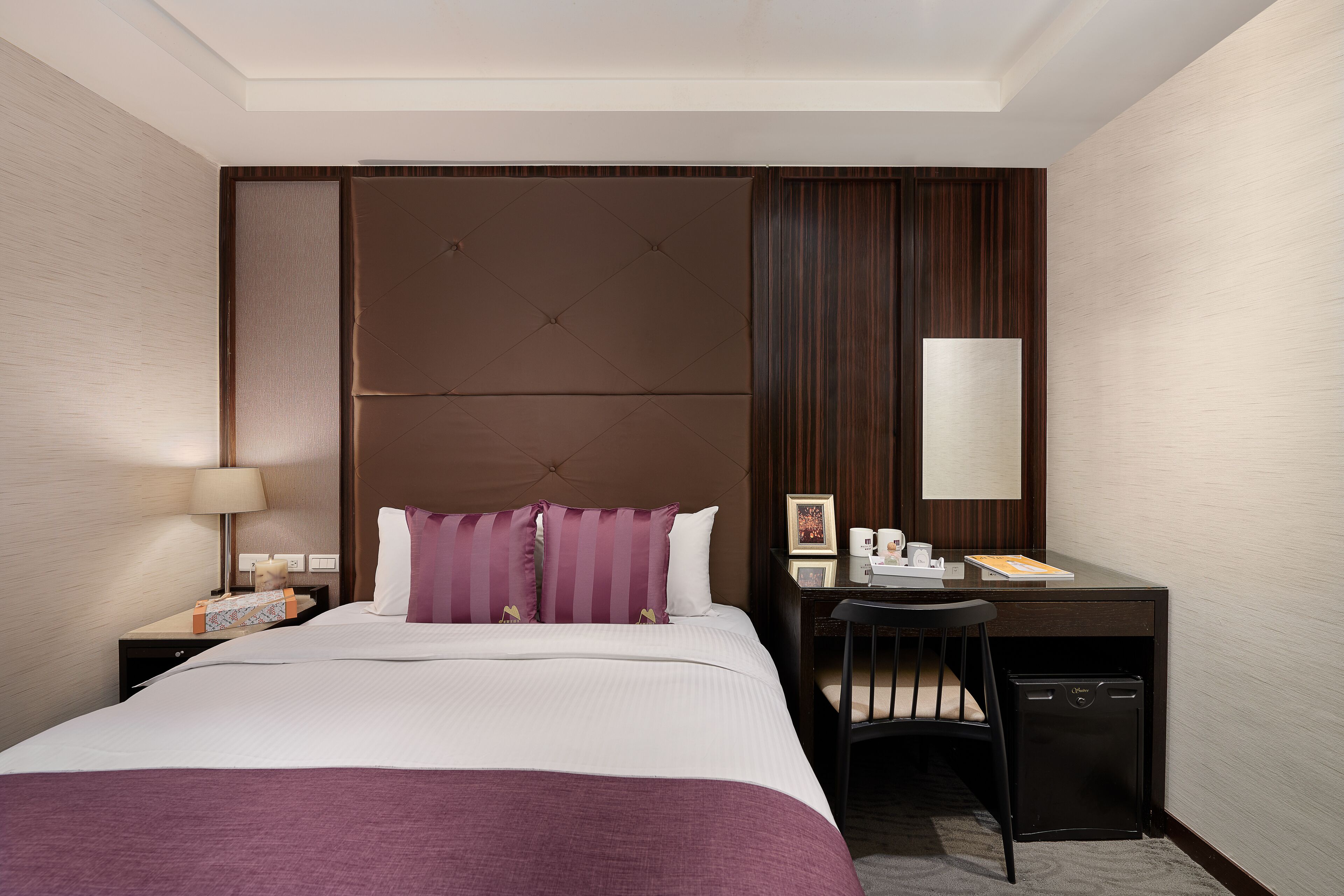 A hotel room with a bed, a desk, a chair, a lamp, and a small cabinet.