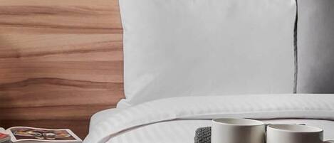 Una cama bien tendida con ropa de cama blanca, una mesita de noche con un libro y una vela, y dos tazas sobre una bandeja.