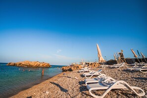 A pebble beach with lounge chairs and umbrellas, a rocky shoreline, and calm sea.