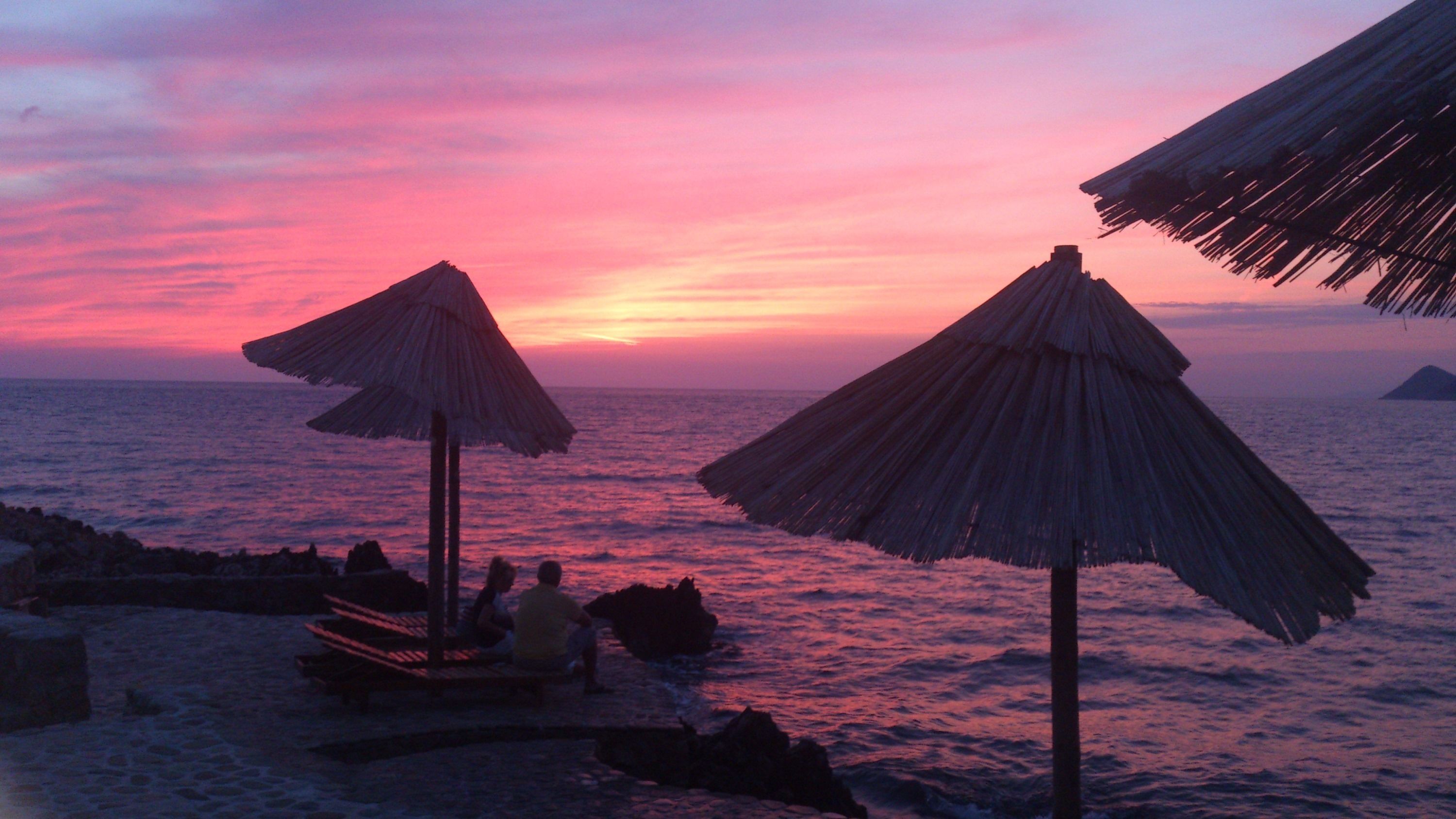A beach scene with two thatched umbrellas and a few people sitting on a bench.