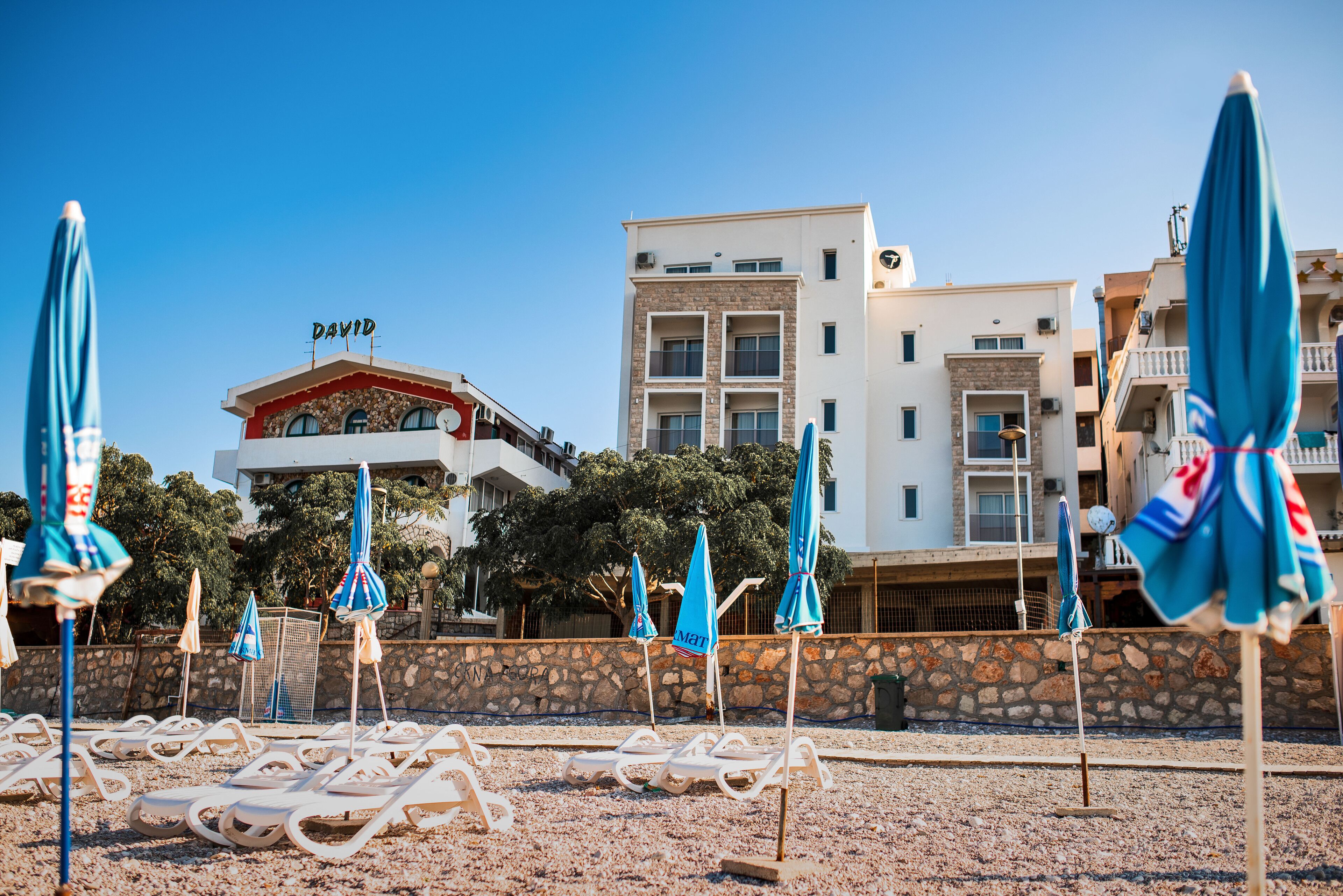 A beachside hotel with lounge chairs and umbrellas, a stone wall, and buildings in the background.