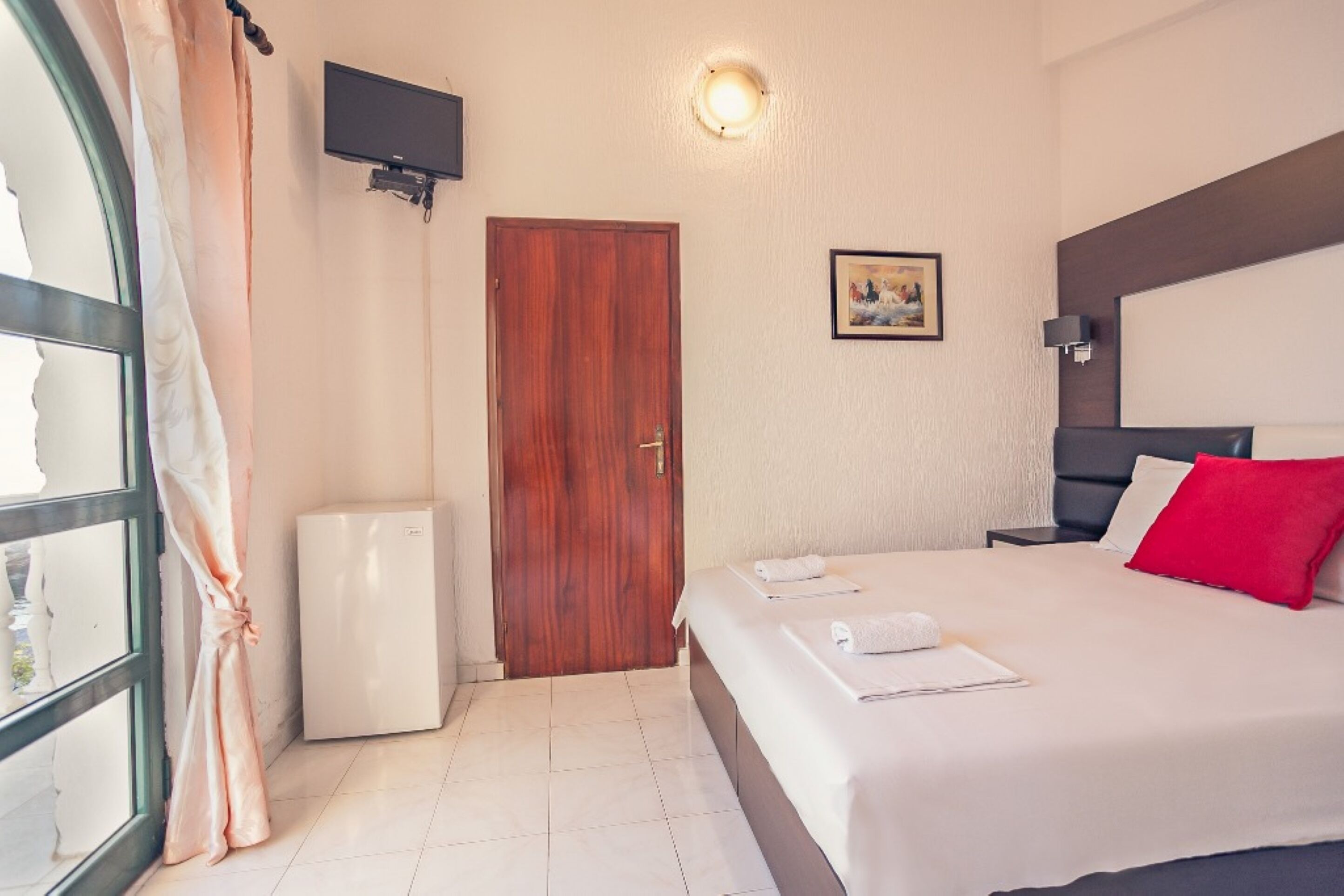 Quarto de hotel com cama, televisão presa à parede, uma porta e um eletrodoméstico branco pequeno.