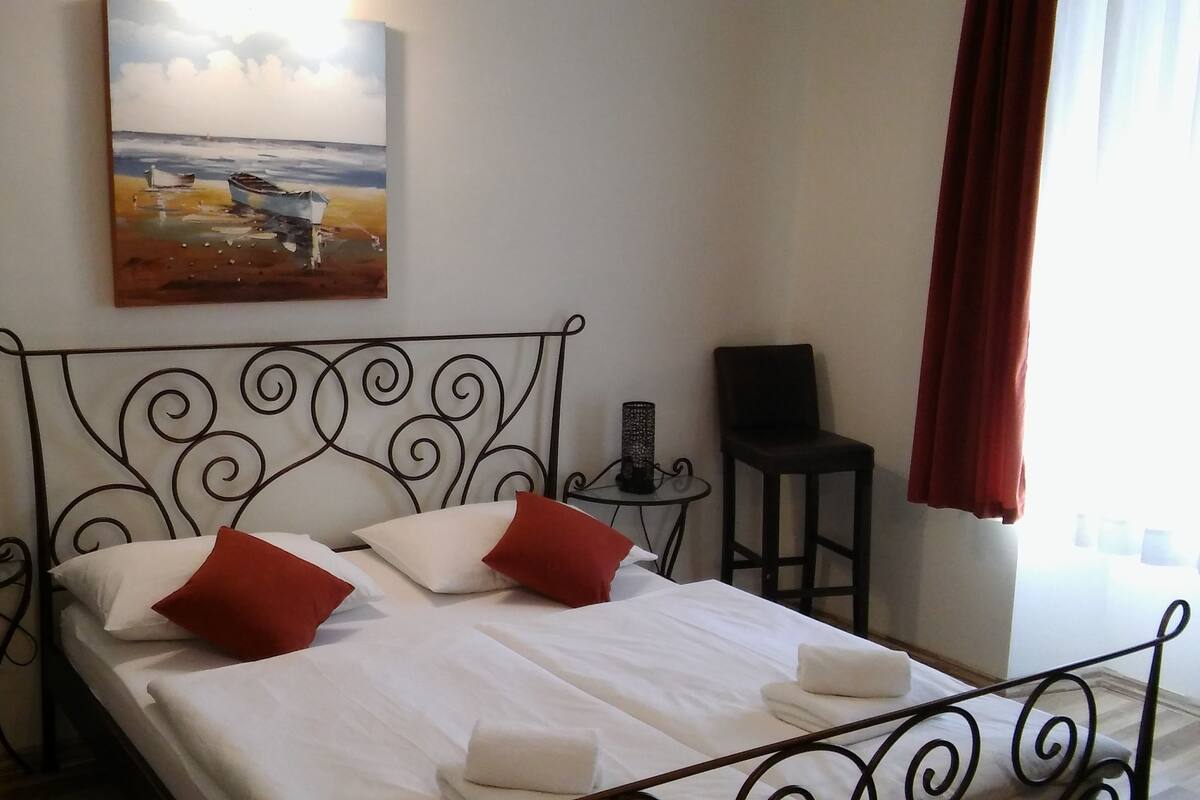 Ein ordentlich bezogenes Bett mit weißen Leinen und roten Kissen, ein Strandbild an der Wand, ein kleiner Tisch mit einer Lampe und ein Sessel.