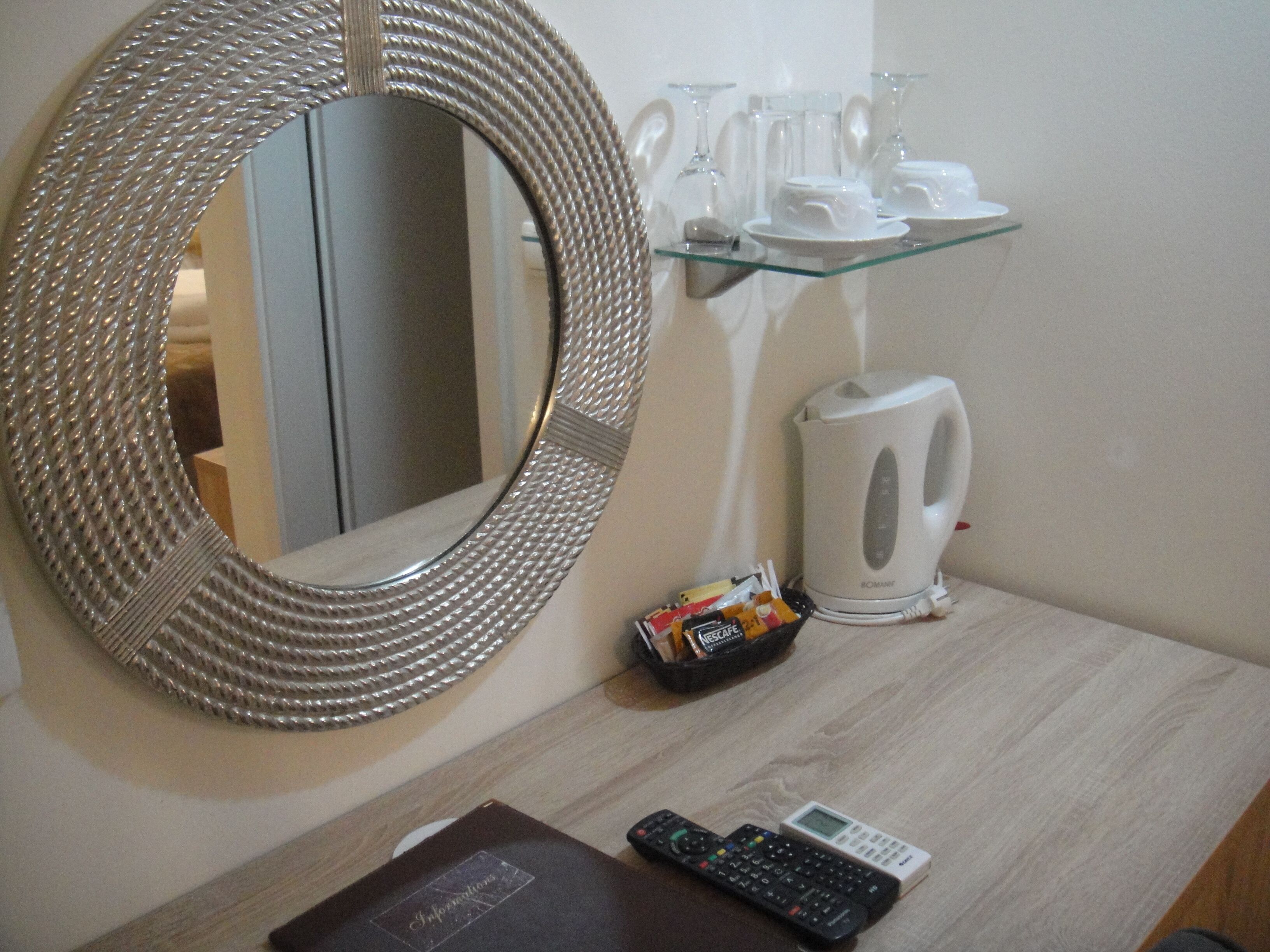 Een badkamer met een ronde spiegel, een glazen plank met toiletartikelen, een waterkoker en een afstandsbediening op het aanrecht.