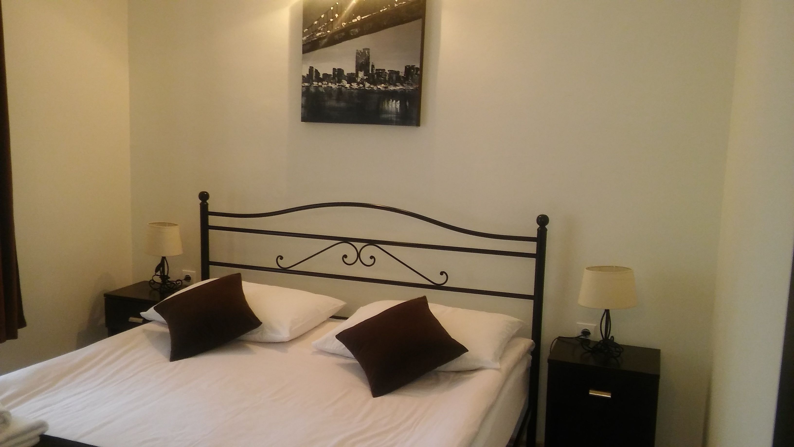 A neatly made bed with white linens and two brown pillows, flanked by two bedside tables with lamps, in a room with a cityscape painting on the wall.