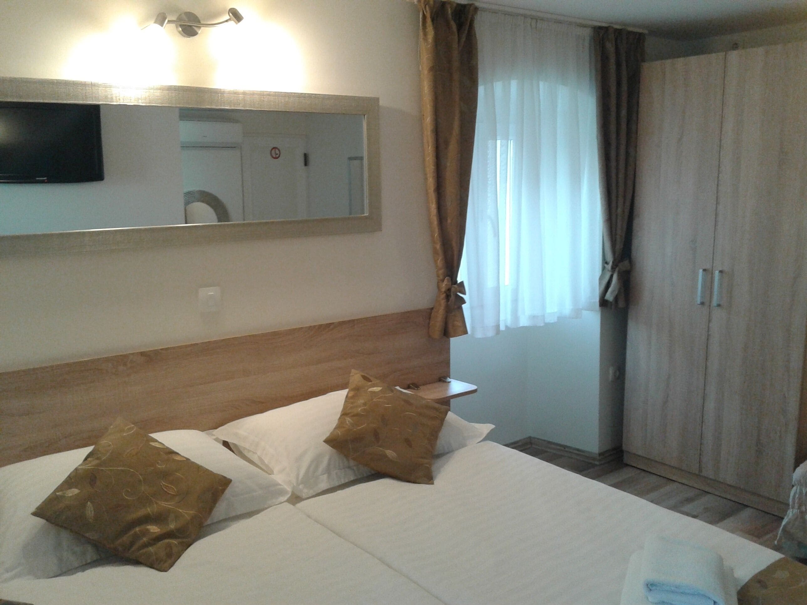 A hotel room with a bed, a television mounted on the wall, a wardrobe, and a window with curtains.