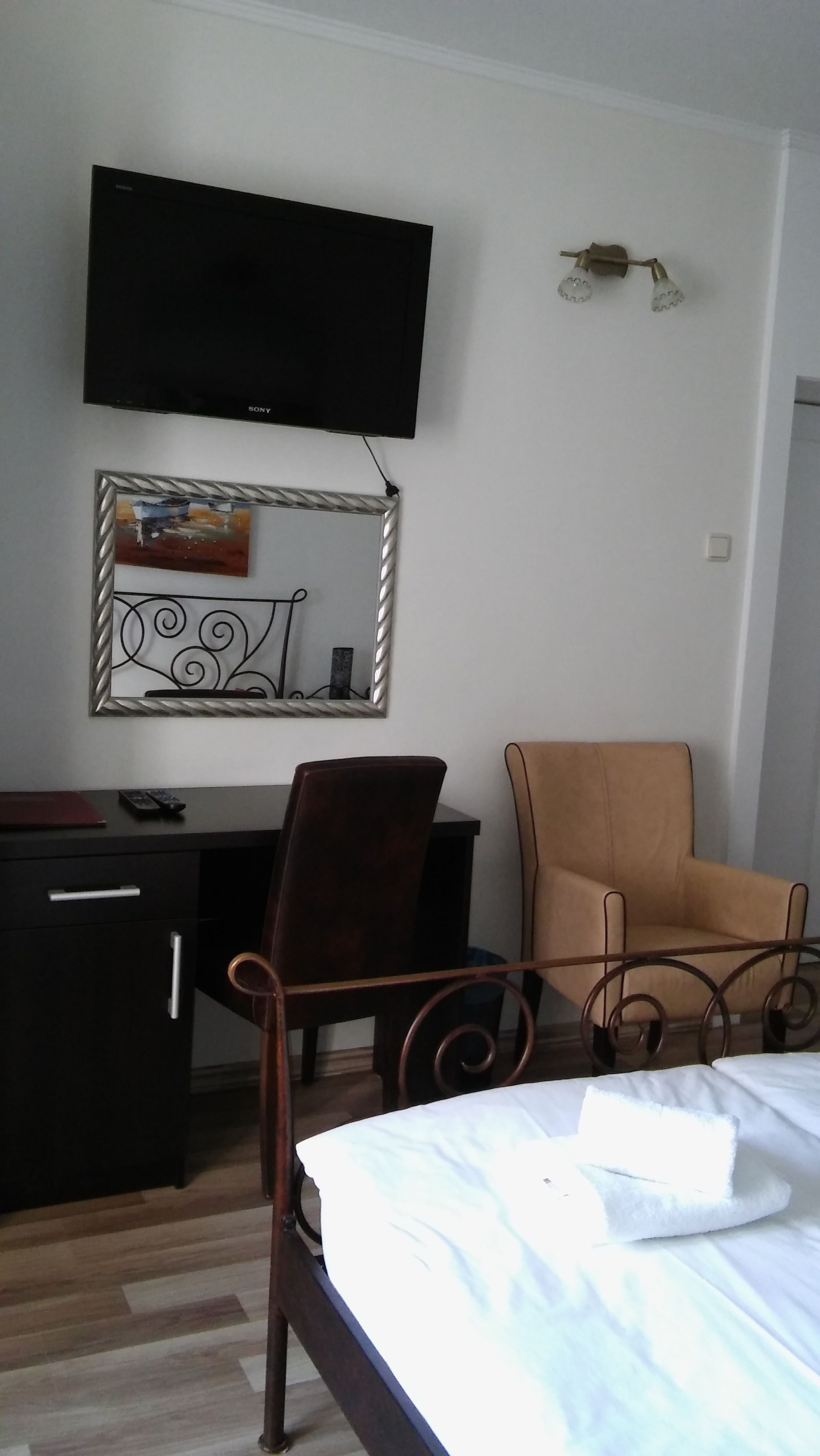 Habitación de hotel con cama, escritorio con silla, televisor montado en la pared y espejo con marco decorativo.