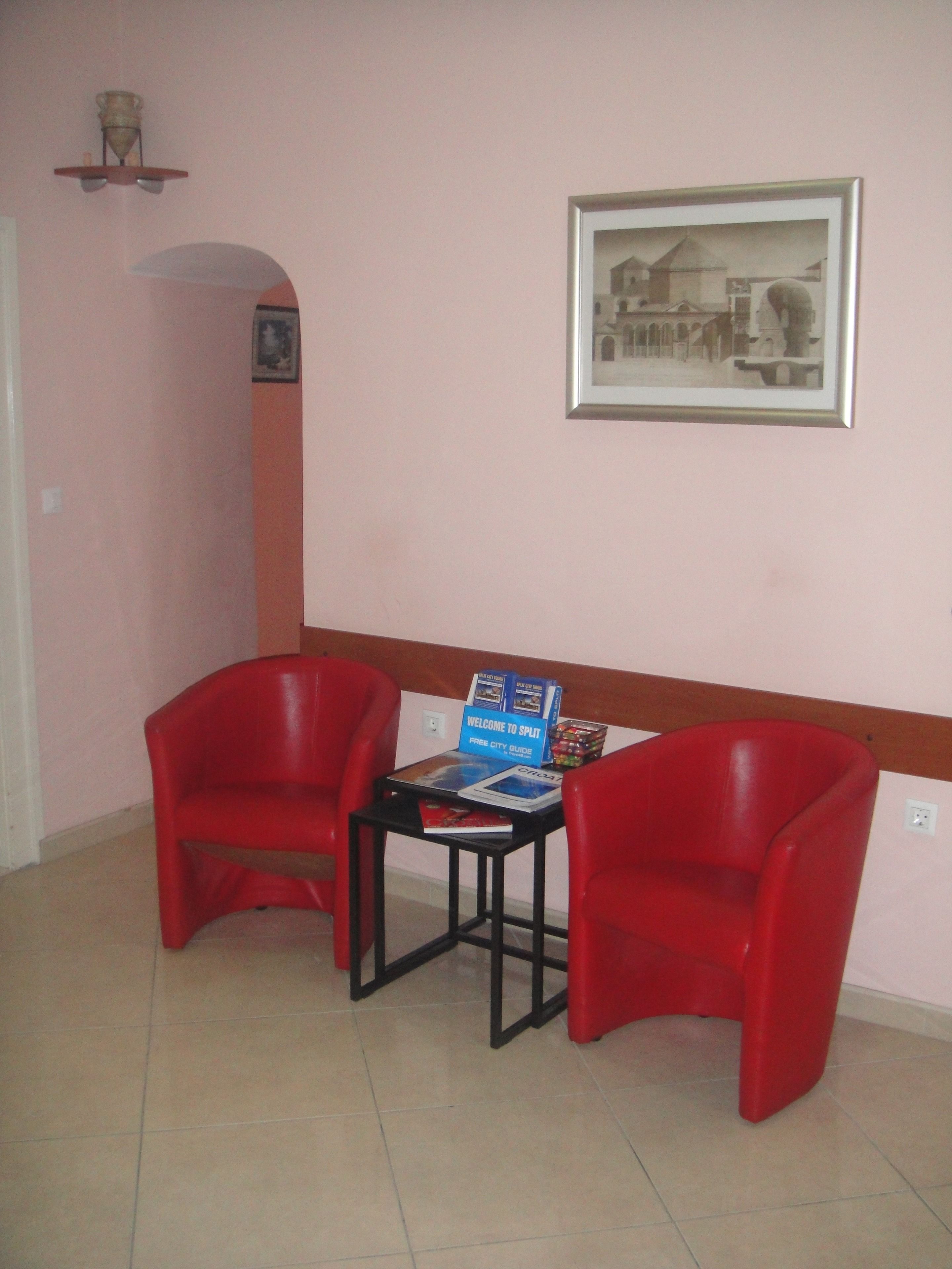 A room with two red chairs, a small table with a welcome sign and a city guide, a framed picture on the wall, and a tiled floor.