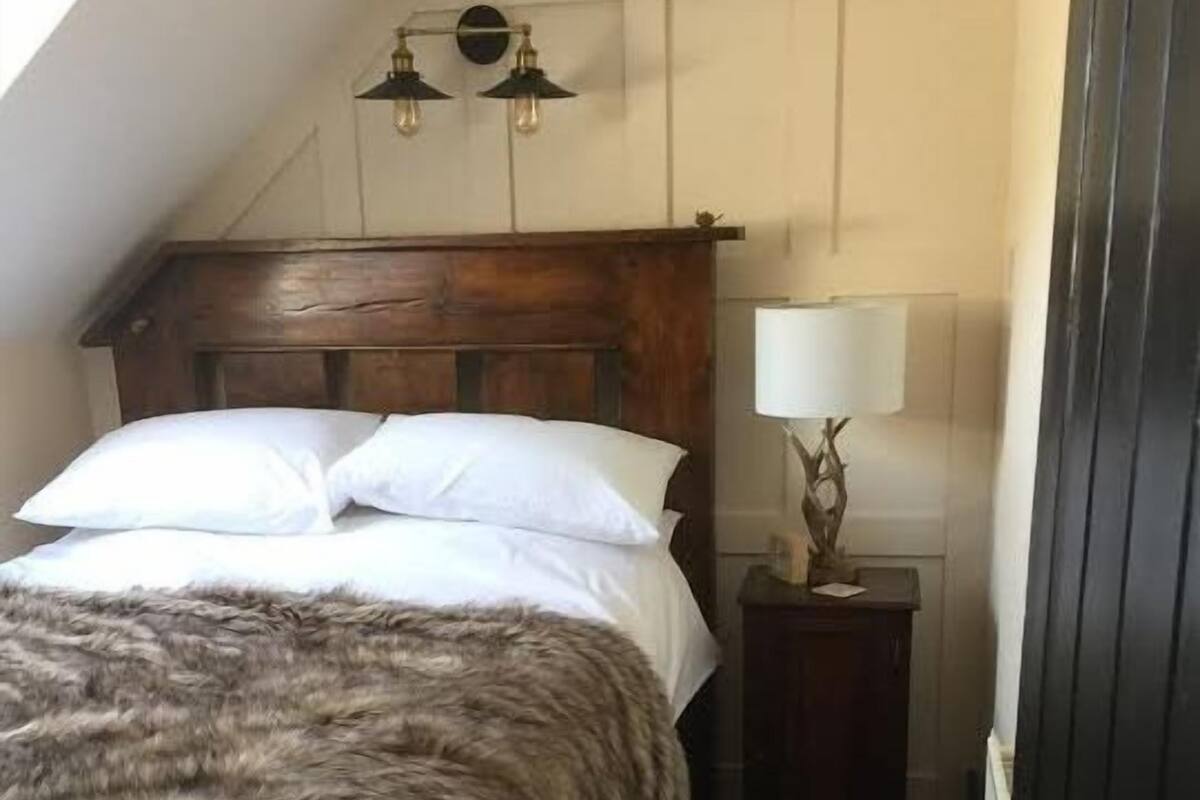 A bedroom with a wooden headboard, two white pillows, a fur throw, a bedside lamp, and a wall-mounted light fixture.