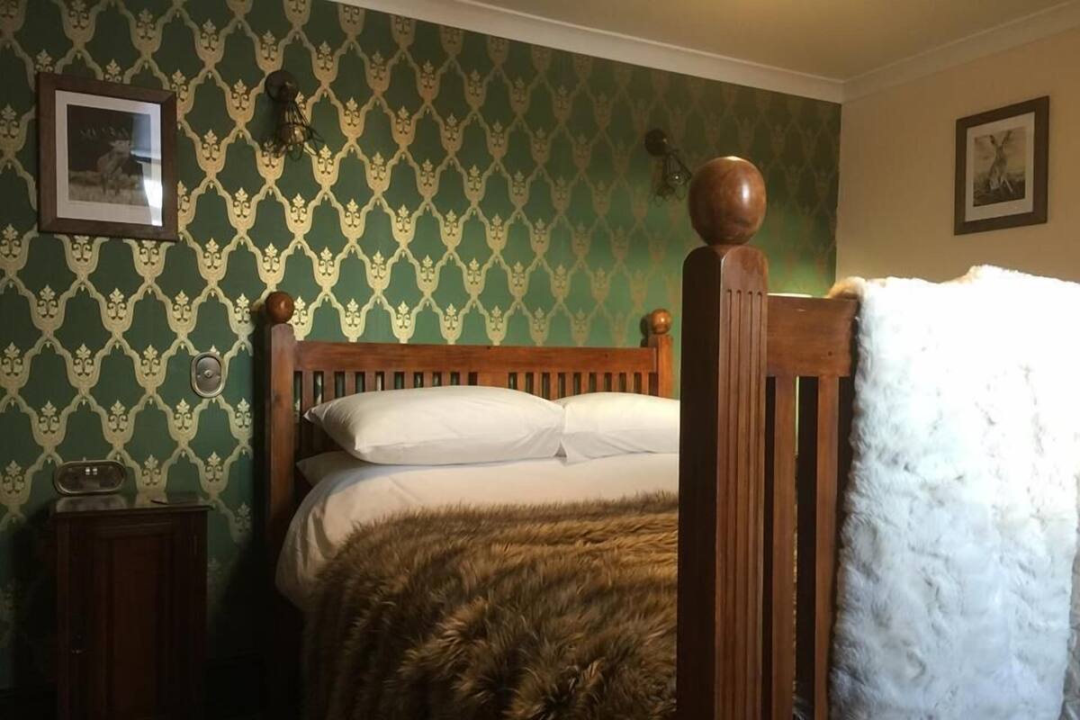 A bedroom with a wooden bed, green patterned wallpaper, a small bedside table, and a fur throw.