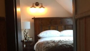 A bedroom with a wooden headboard, a bed with white pillows, a nightstand with a lamp, and a wall-mounted light fixture. - Balsporran Bed And Breakfast (Dalwhinnie)