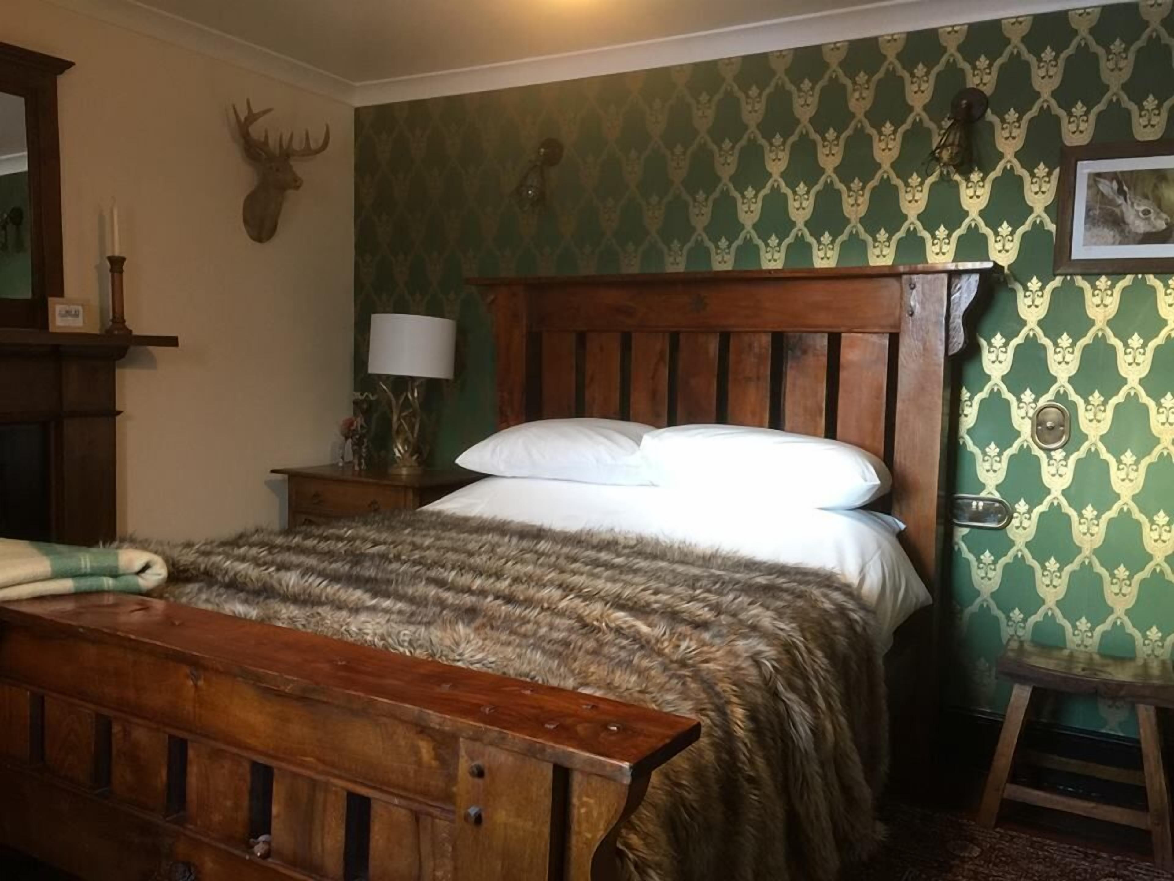 Quarto com cama de madeira, mesa de cabeceira com abajur, cabeça de veado fixada na parede e papel de parede estampado.