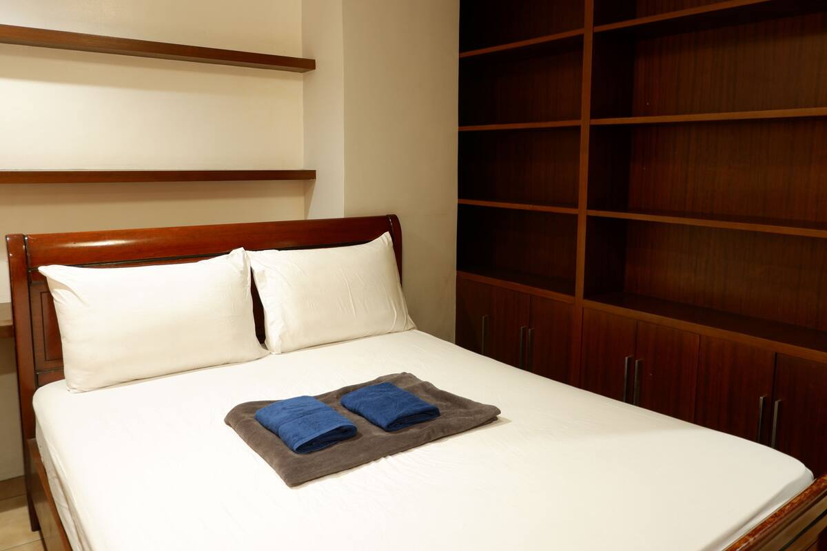 A neatly made bed with white pillows and a gray blanket, wooden headboard, and a large wooden wardrobe.