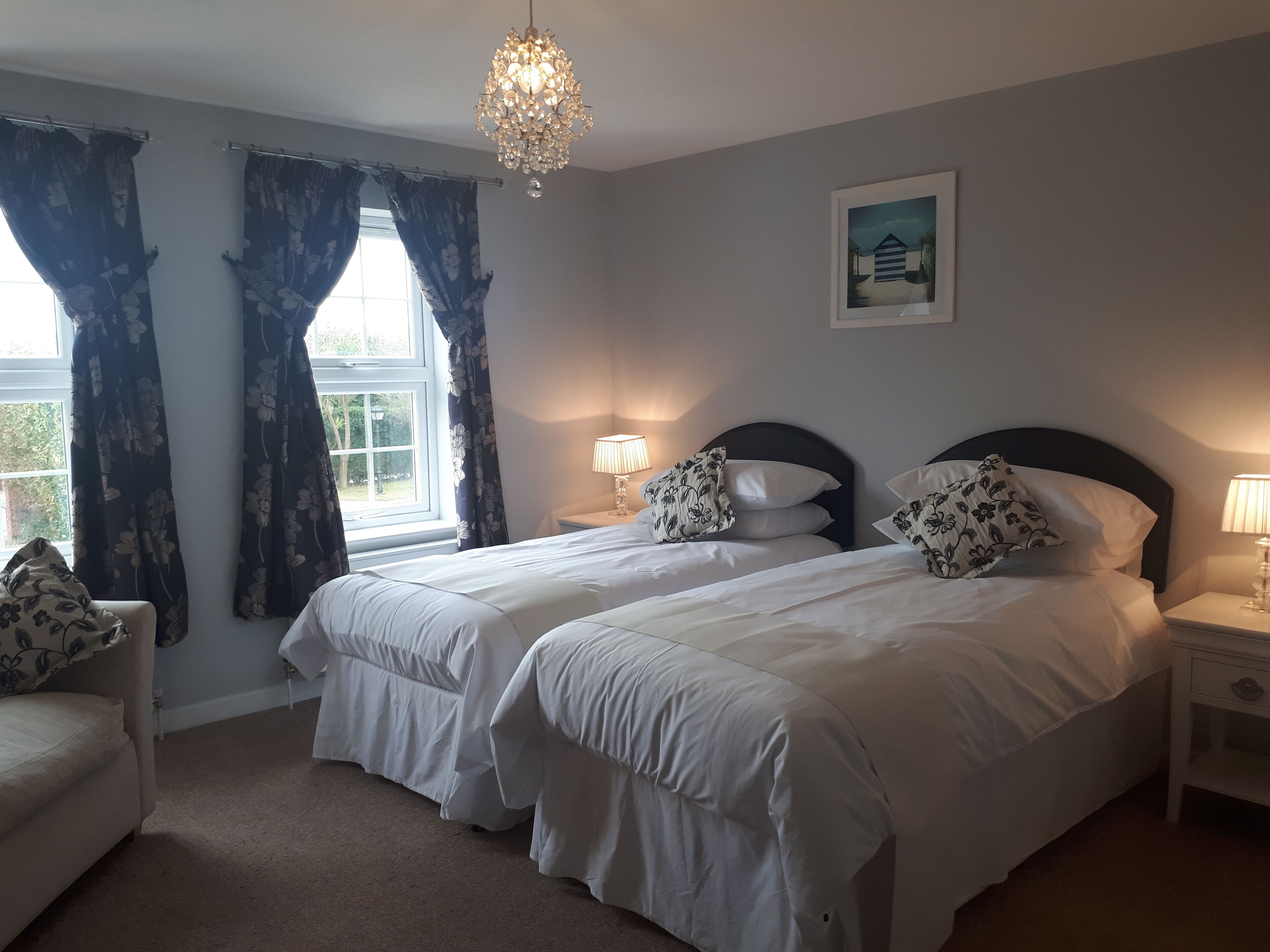 A bedroom with two beds, a chandelier, a window with curtains, a bedside table with a lamp, and a painting on the wall.