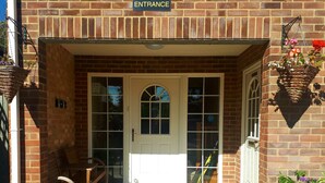 A brick building entrance with a white door, a bench, and hanging flower baskets.