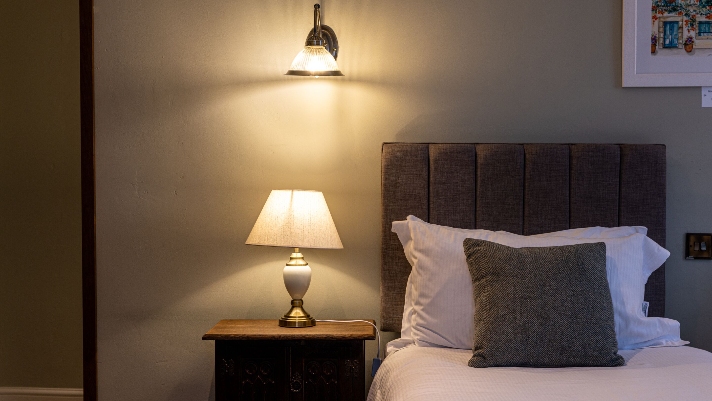 A neatly made bed with white linens and a grey headboard, a bedside lamp, and a wall-mounted light fixture.