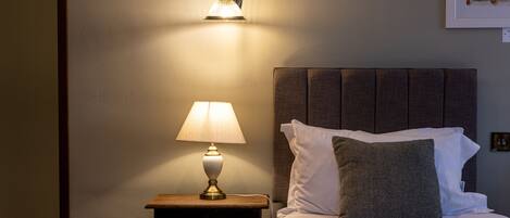 A neatly made bed with white linens and a grey headboard, a bedside lamp, and a wall-mounted light fixture.