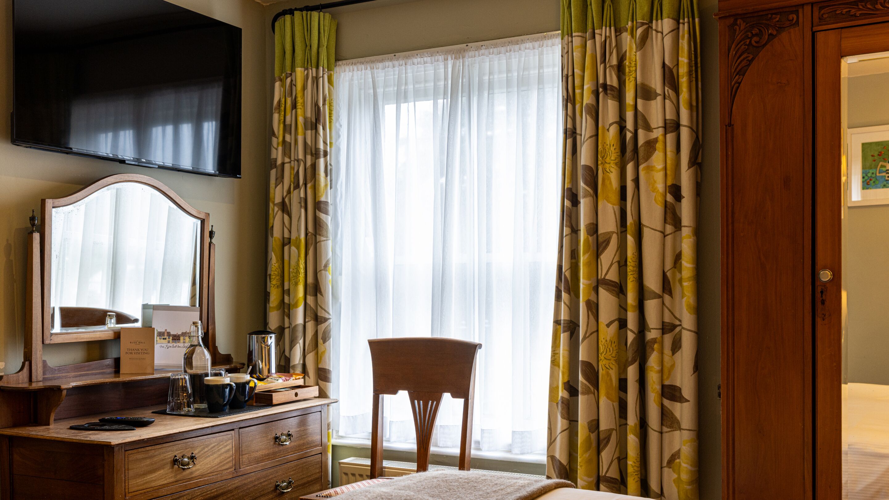 A hotel room with a wooden dresser, a mirror, a television mounted on the wall, a chair, and a large window with sheer curtains.