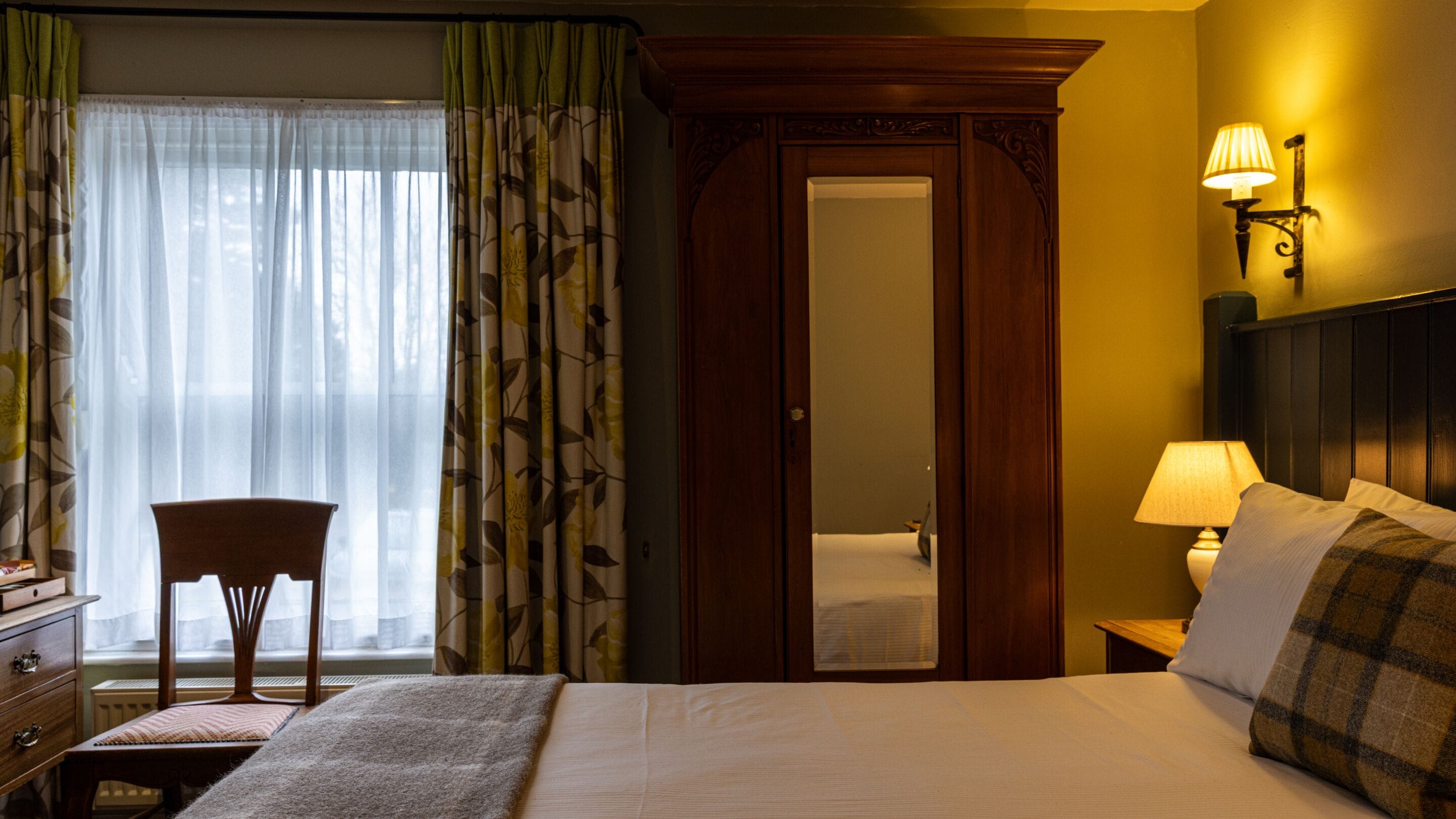 Habitación de hotel con cama, una silla, mesita de noche con lámpara, armario y ventana con cortinas.