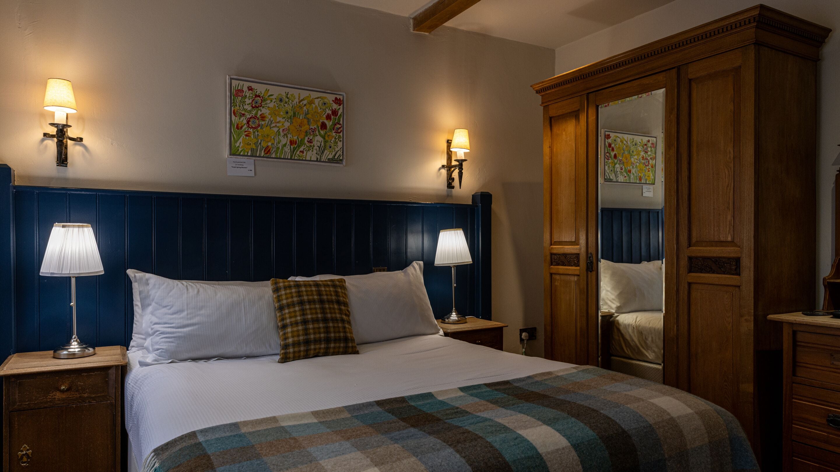 A neatly made bed with a plaid blanket, two bedside lamps, a wooden wardrobe, and floral artwork on the walls.