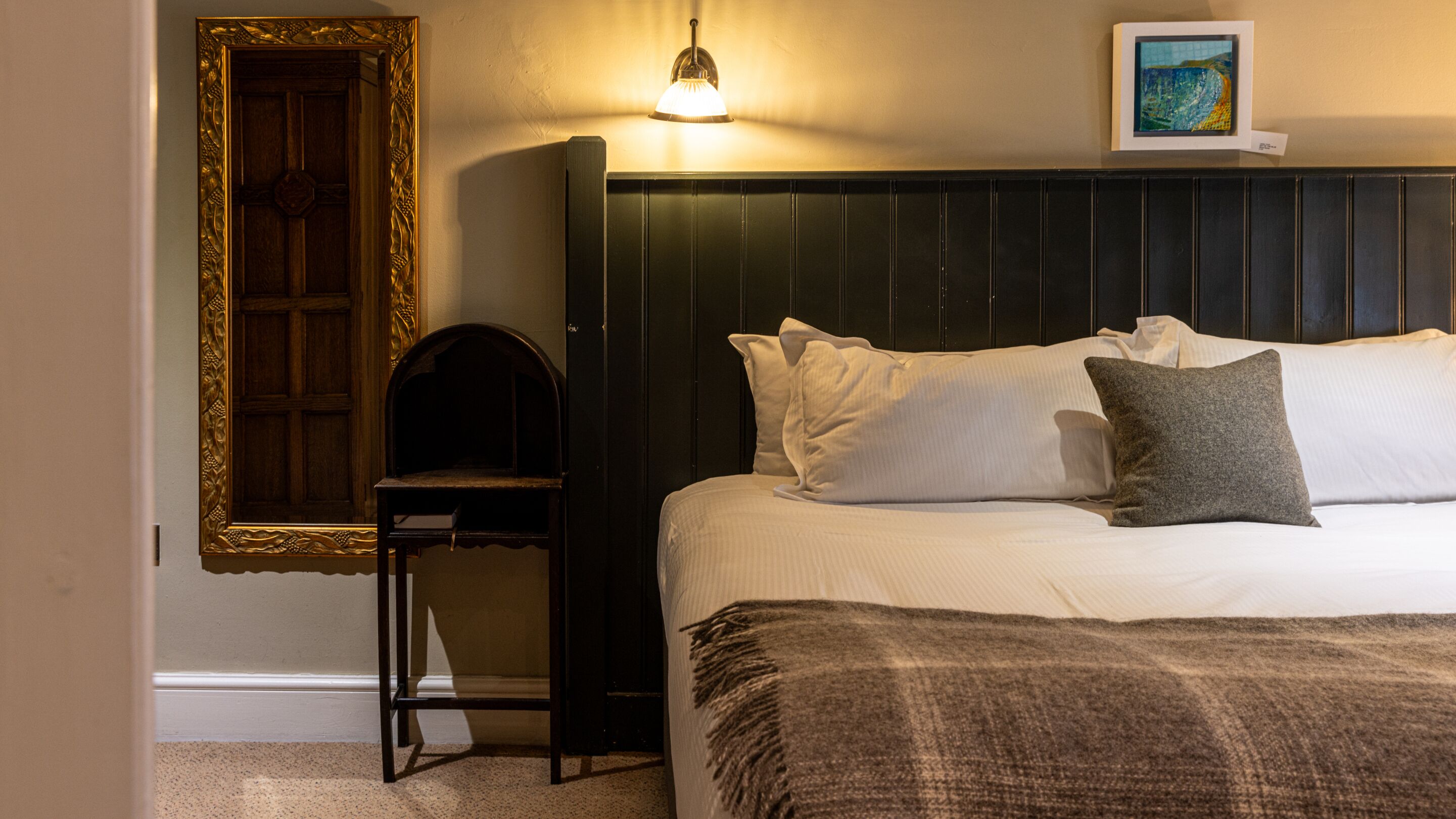 A bedroom with a bed, pillows, a bedside table, a lamp, a framed picture, and a mirror.