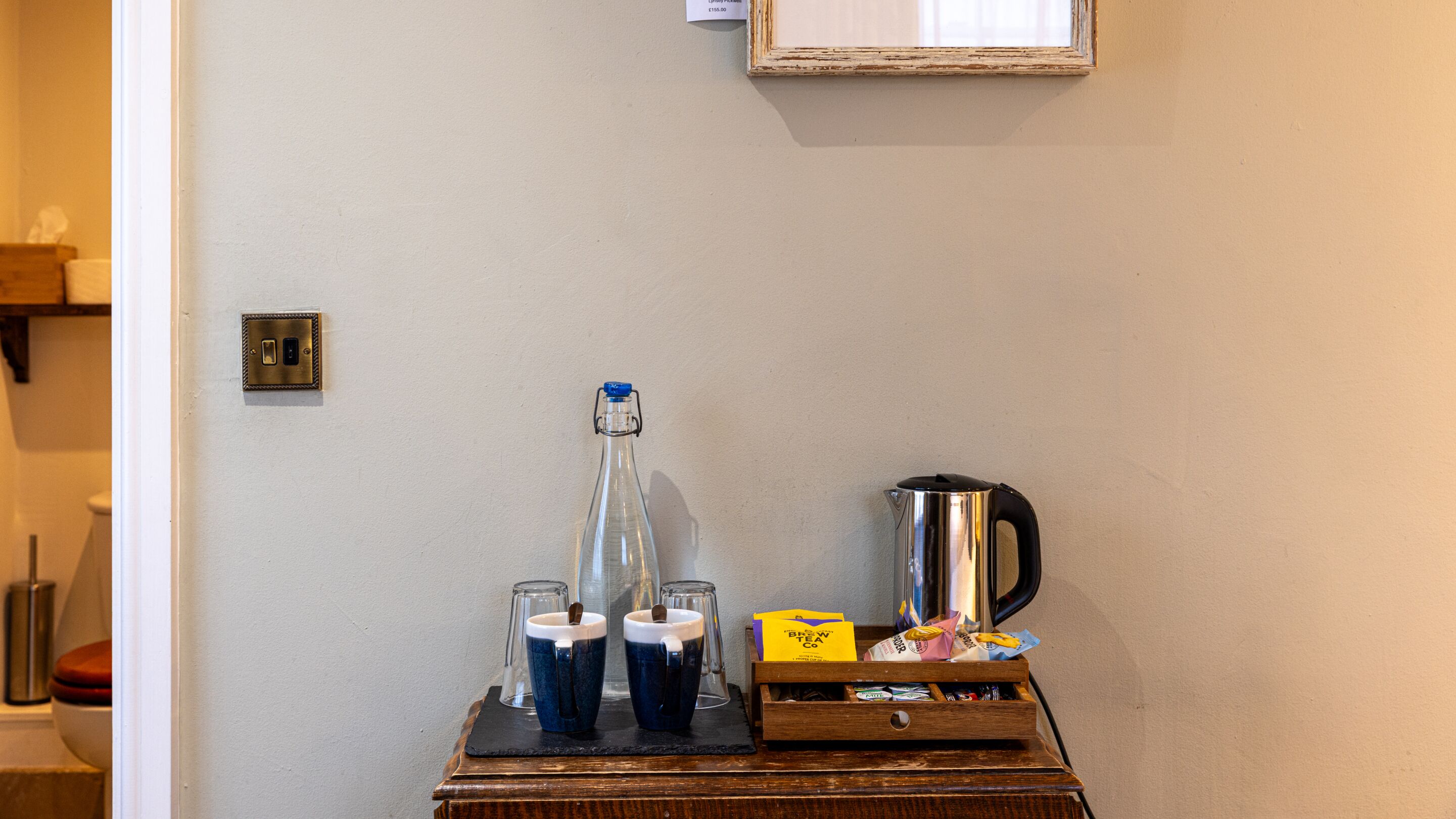 A room with a wooden side table holding a kettle, two mugs, a glass carafe, and a box of tea.