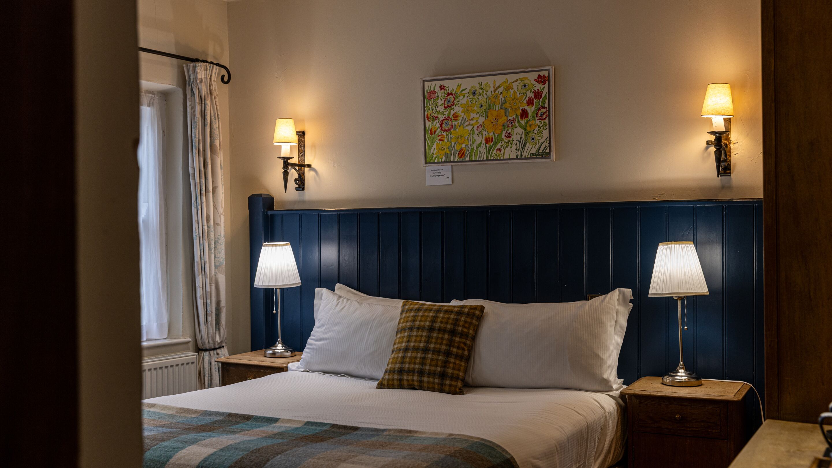 A hotel room with a large bed, two bedside tables with lamps, a window with curtains, and a floral painting on the wall.