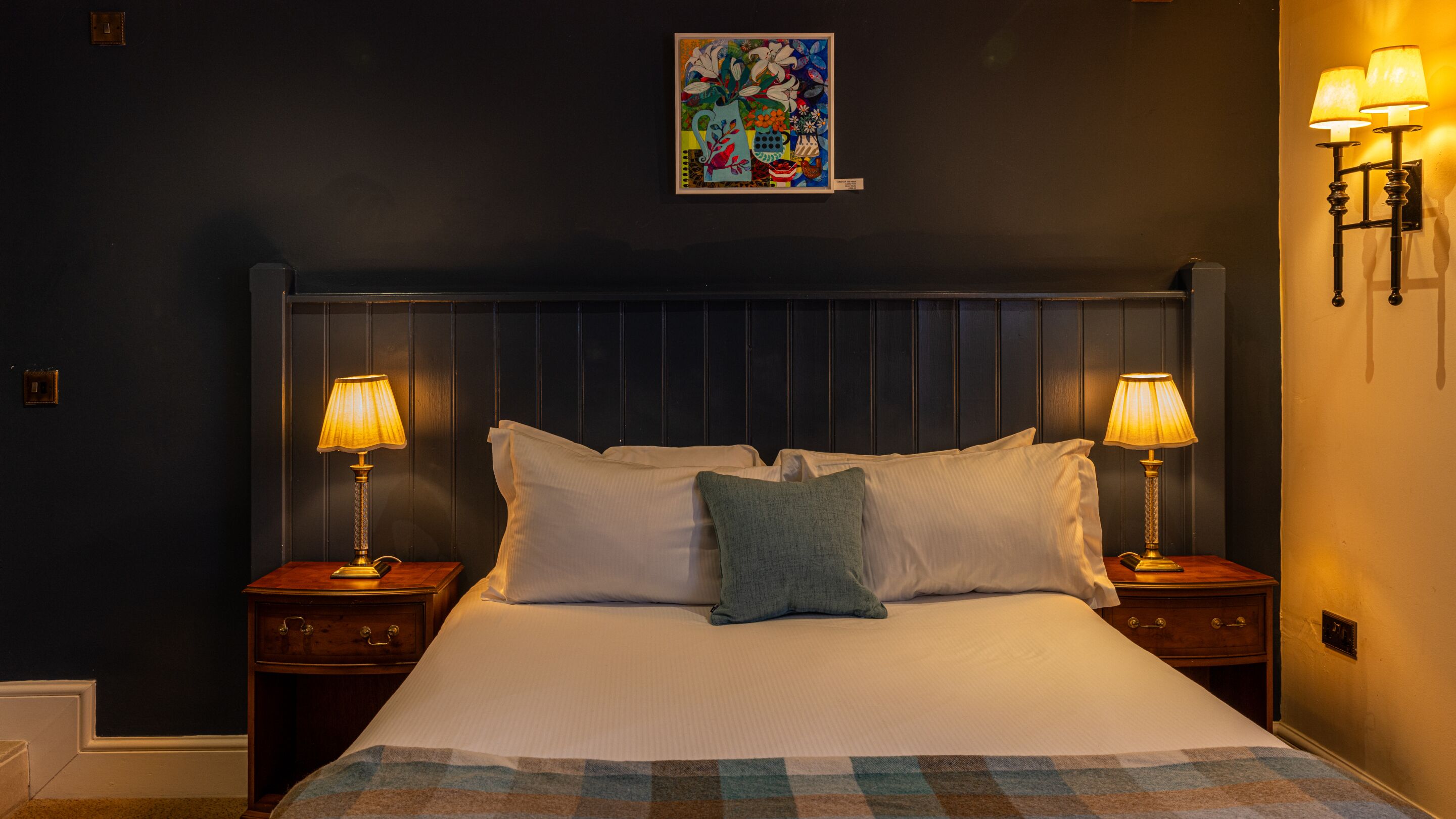A hotel room with a large bed, two bedside lamps, a painting on the wall, and a checkered throw on the bed.