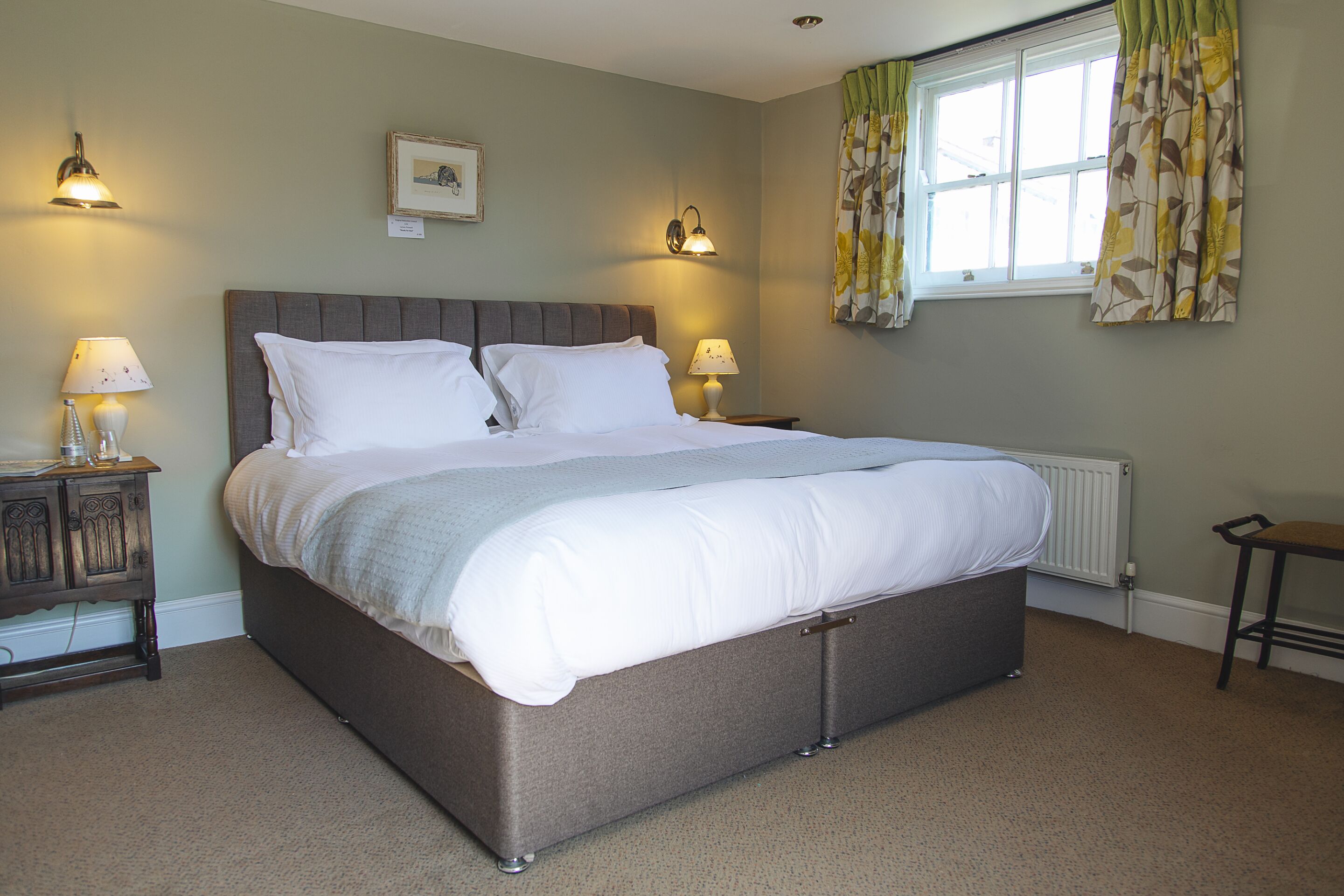 A neatly made bed with a grey headboard, two bedside lamps, and a window with curtains.