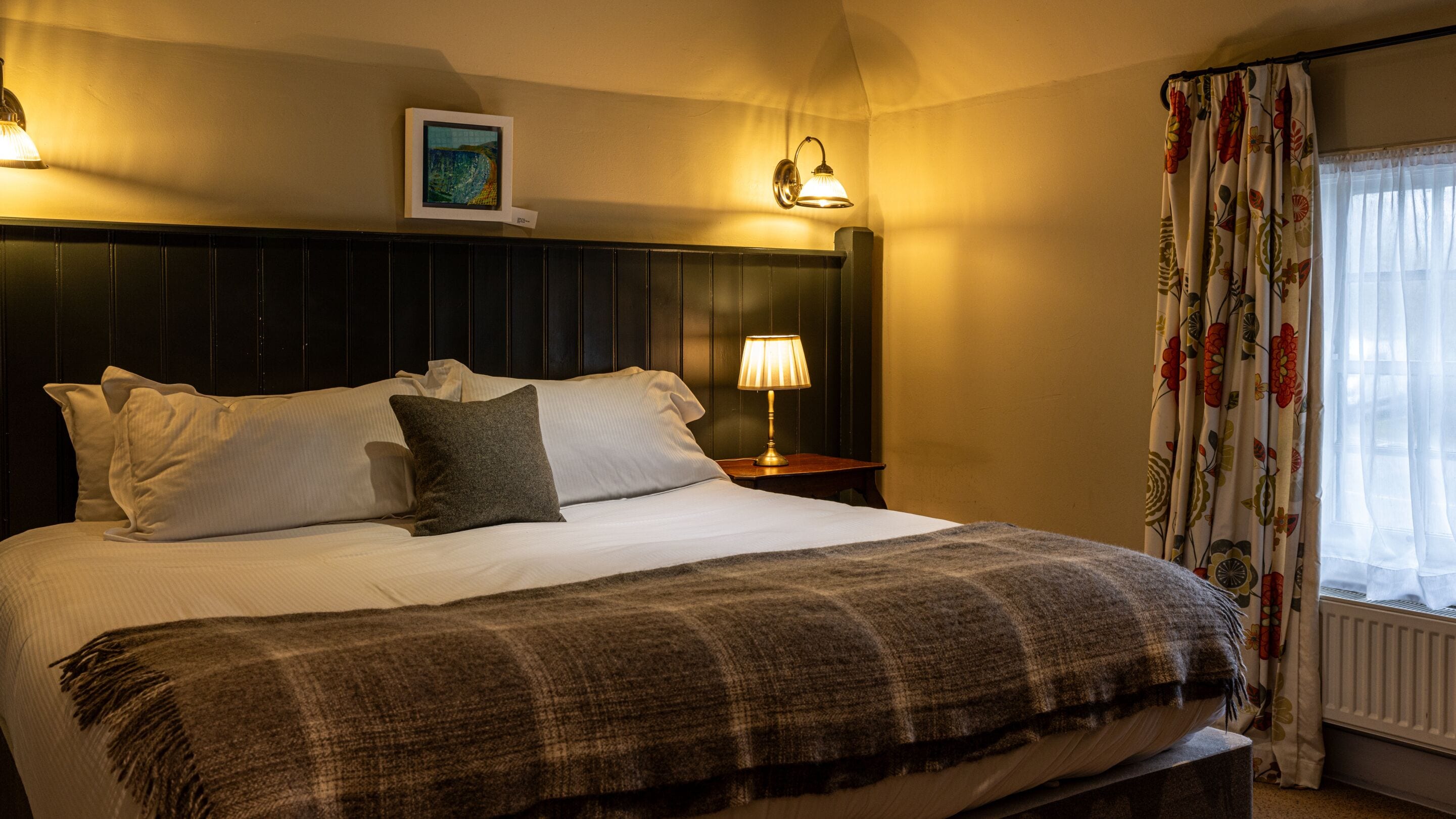 Un lit bien fait, avec du linge de lit blanc et un plaid marron, une lampe de chevet et un tableau encadré accroché au-dessus de la tête de lit.