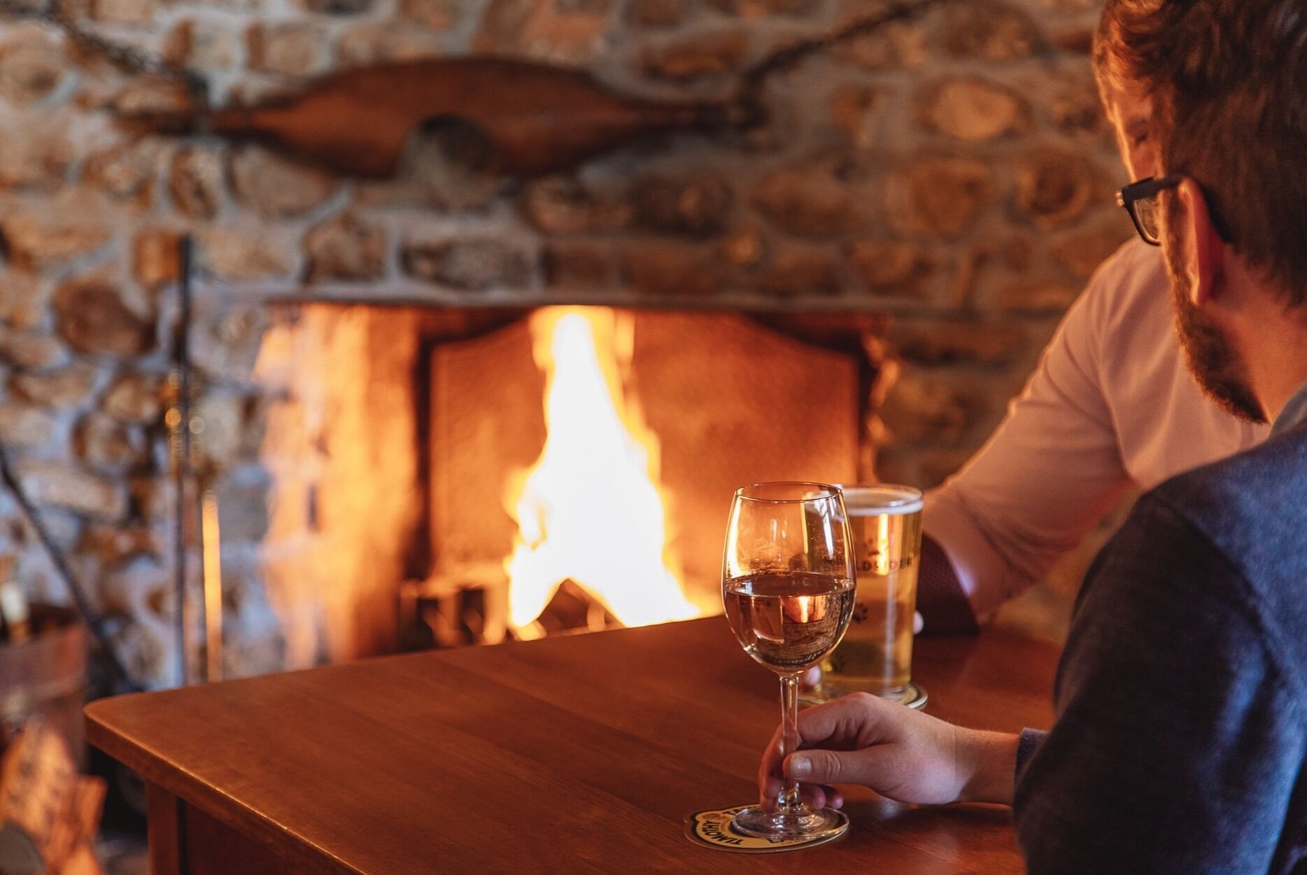Une personne tenant un verre de vin à une table en bois, avec une cheminée en pierre en arrière-plan.